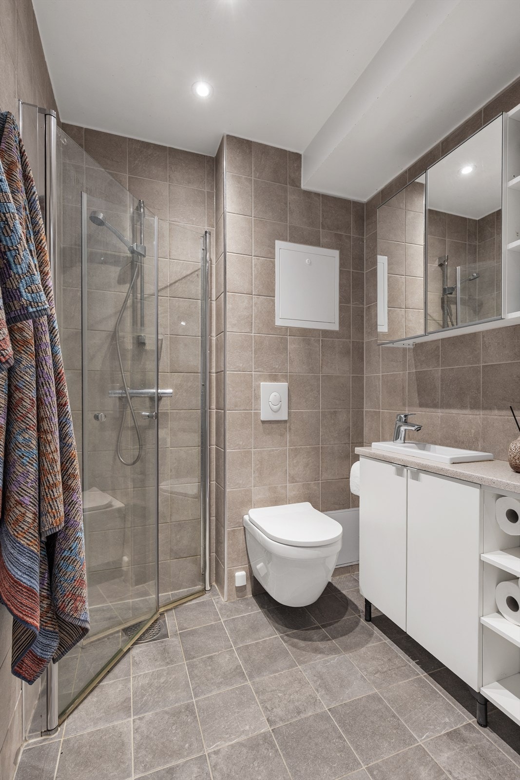 Toalettet er pent flislagt på gulv og vegger med elektrisk varme i gulvet. Badet er innredet med vegghengt klosett, servant med innredning, speilskap og dusjhjørne med innfellbare glassdører. Galleribilde