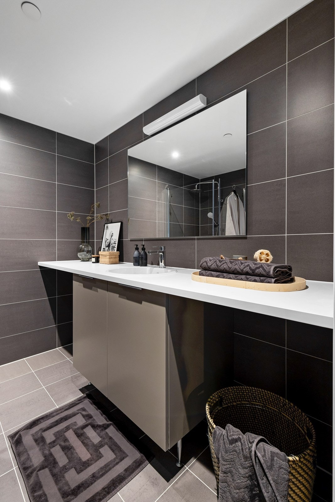 Badet er ellers innredet med vegghengt toalett, servantinnredning med skap, heldekkende benkeplate, porselenservant med ettgreps armatur, vegghengt speil og lys. Opplegg og plass for vaskemaskin under benkeplate. Galleribilde