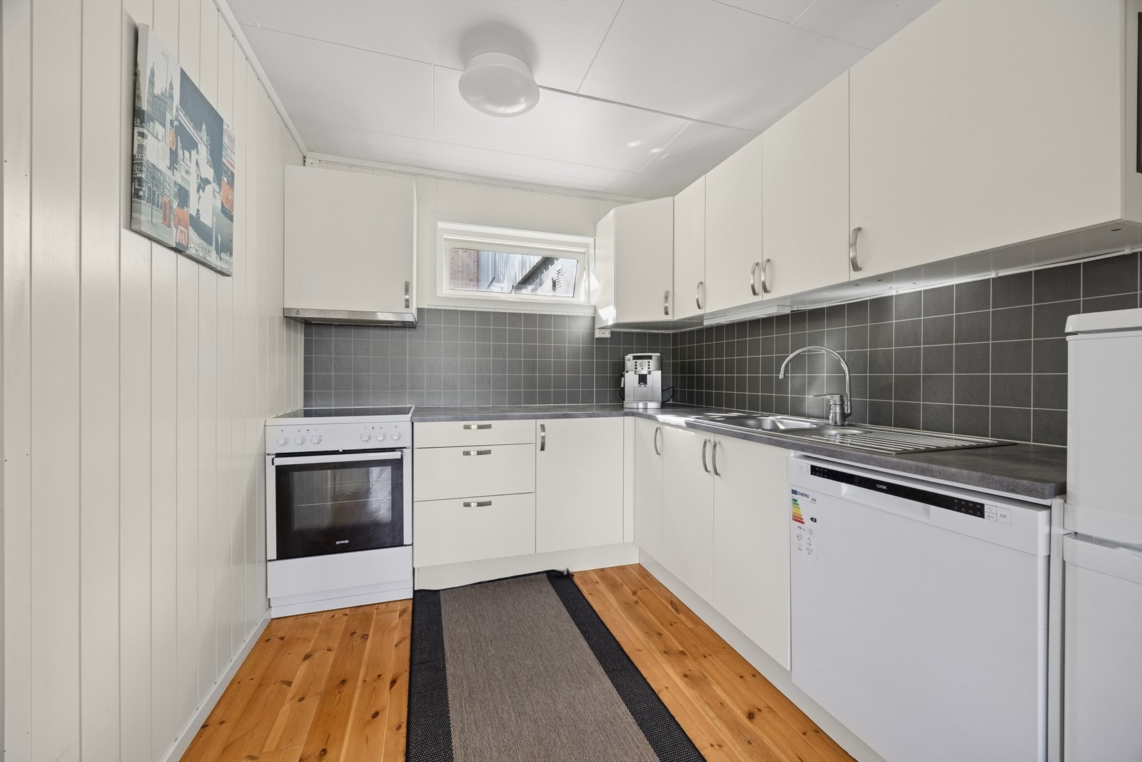 Her finner du alt du trenger i hverdagen - komfyr med platetopp, oppvaskmaskin samt kjøleskap med frysedel. En effektiv og innbydende kjøkkenløsning som passer perfekt til sokkelleilighetens behov. Galleribilde