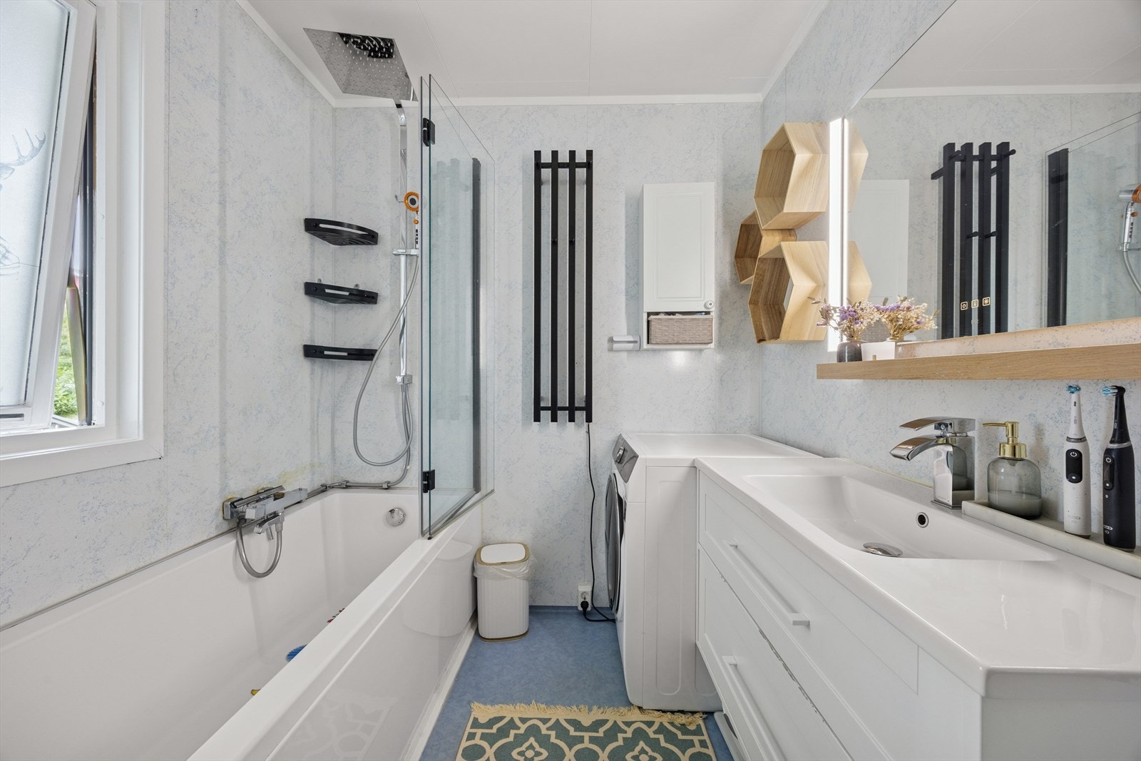 Badet er innredet med belegg på gulv og våtromsplater på vegger. Servant med innredning, speil med belysning og toalett. Rommet er også utstyrt med badekar med glassvegg og dusjopplegg samt plass til vaskemaskin. Galleribilde