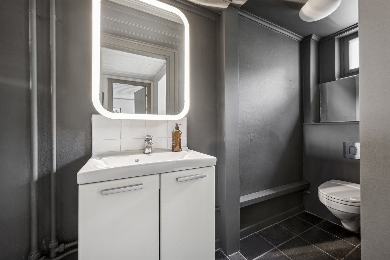 Toalettrom med fliser på gulv og malt puss på vegger. Innredning med nedfelt servant. Veggmontert toalett. Galleribilde