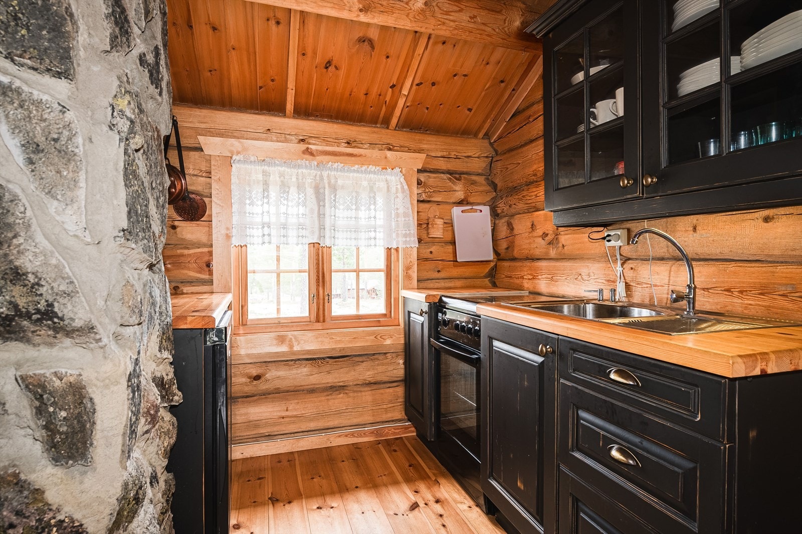 Pent hyttekjøkken med sortbeiset innredning. Vanntilførsel til servant fra vannbeholder tilknyttet 12V vannpumpe, avløp med gråvann til terreng. Galleribilde