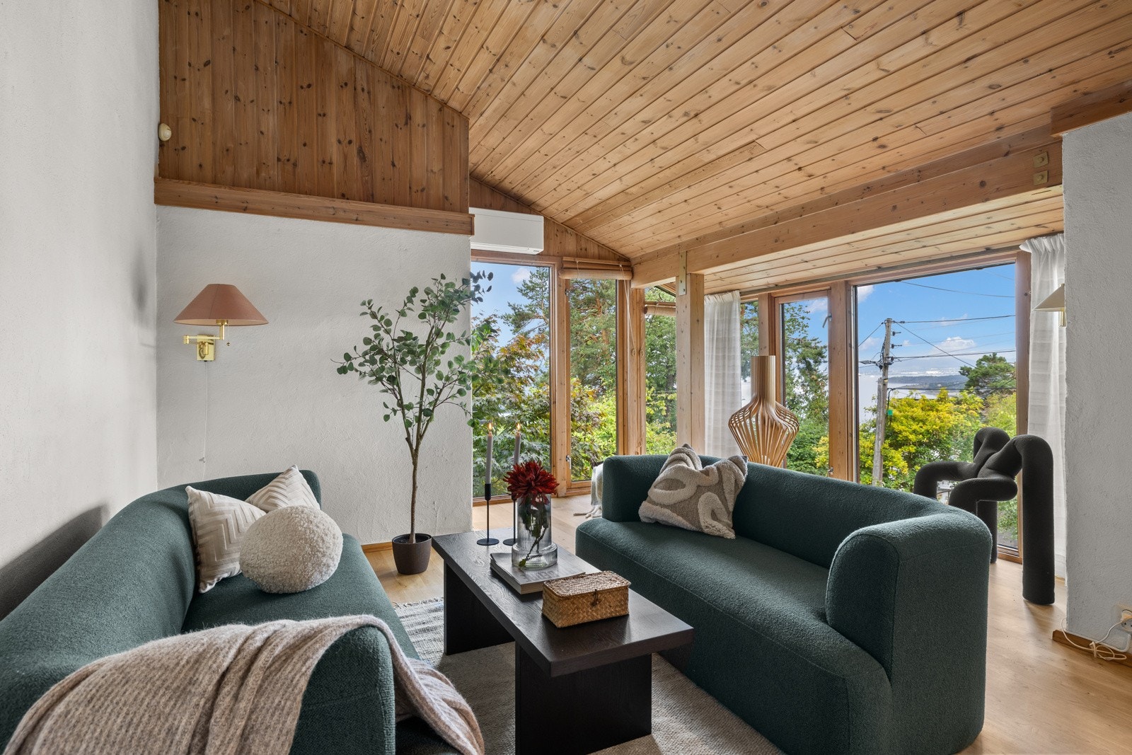 Boligen har en romslig stue med trepanel i tak og vegger, og utsikt mot grønne omgivelser og fjorden. Gulv-til-tak-vinduene slipper inn rikelig med naturlig lys. Galleribilde