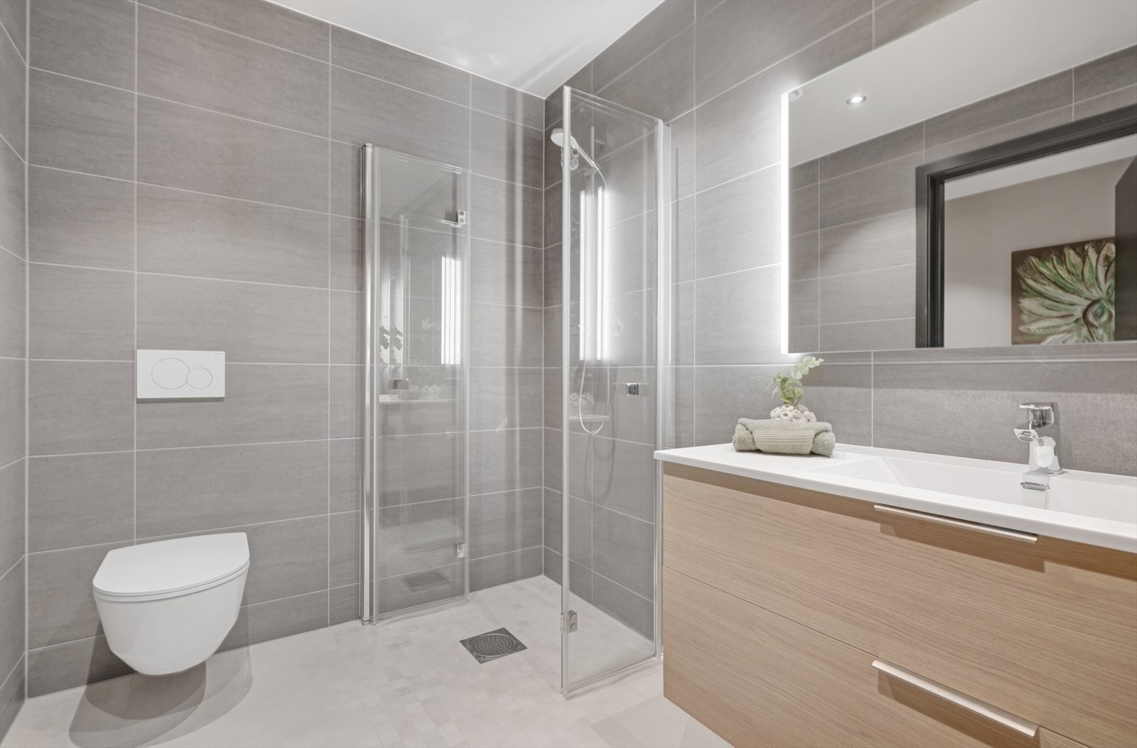 Lekkert og moderne flislagt bad med vegghengt toalett, dusjhjørne og servant med speil, belysning og innredning. Galleribilde