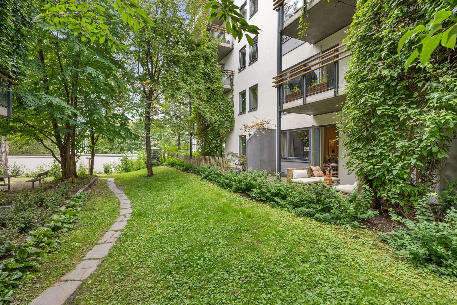 Uteplassen vender ut mot felles hageareal med trær, busker, og frodige klatreplanter som svinger seg opp husveggen. Galleribilde