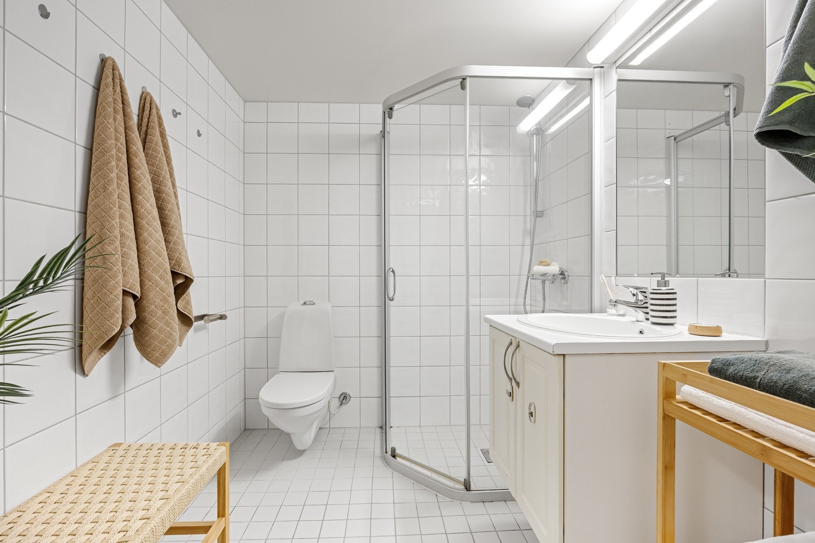 Badet har en hvit servantinnredning, speil med belysning, veggmontert toalett og et dusjhjørne med skyvedører. Galleribilde