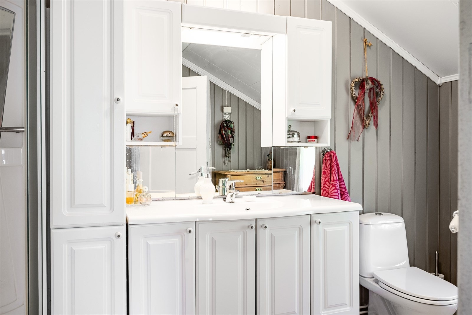 Lys baderomsinnredning med profilerte dører og heldekkende servant, speil med belysning og vegghengte skap over. Galleribilde