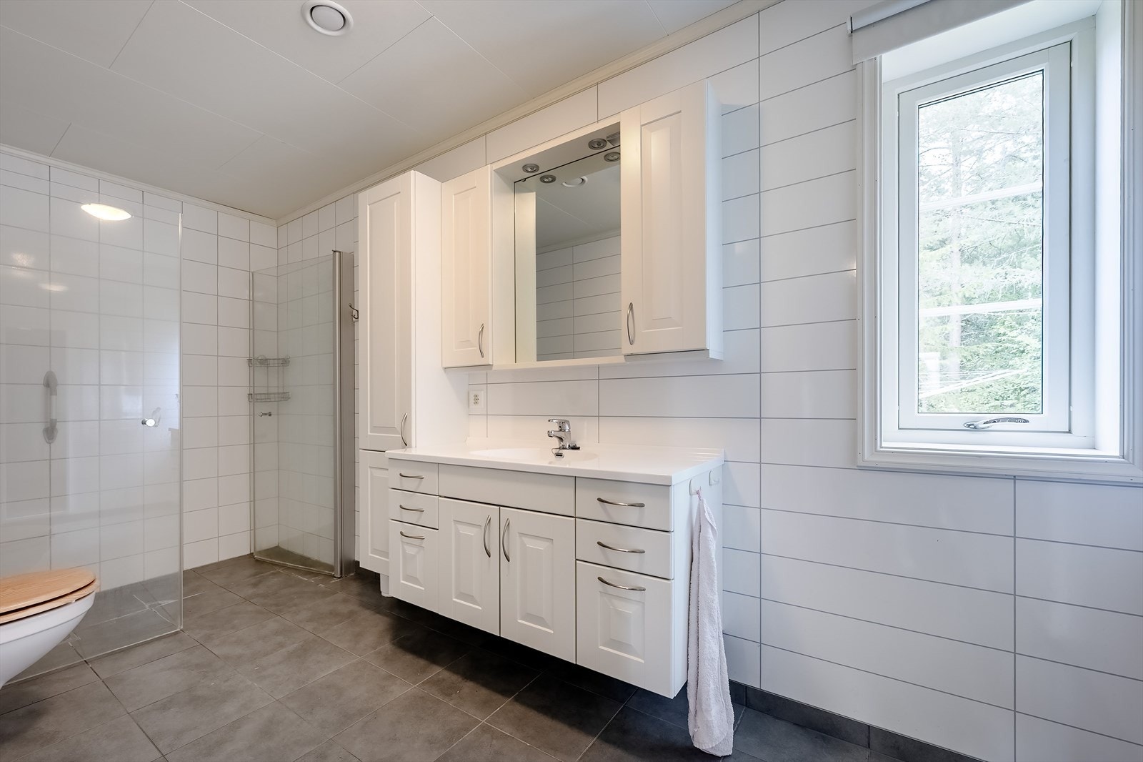 Badet har flislagt gulv med undervarme, stor dusjnisje og innredning med god skapplass Galleribilde