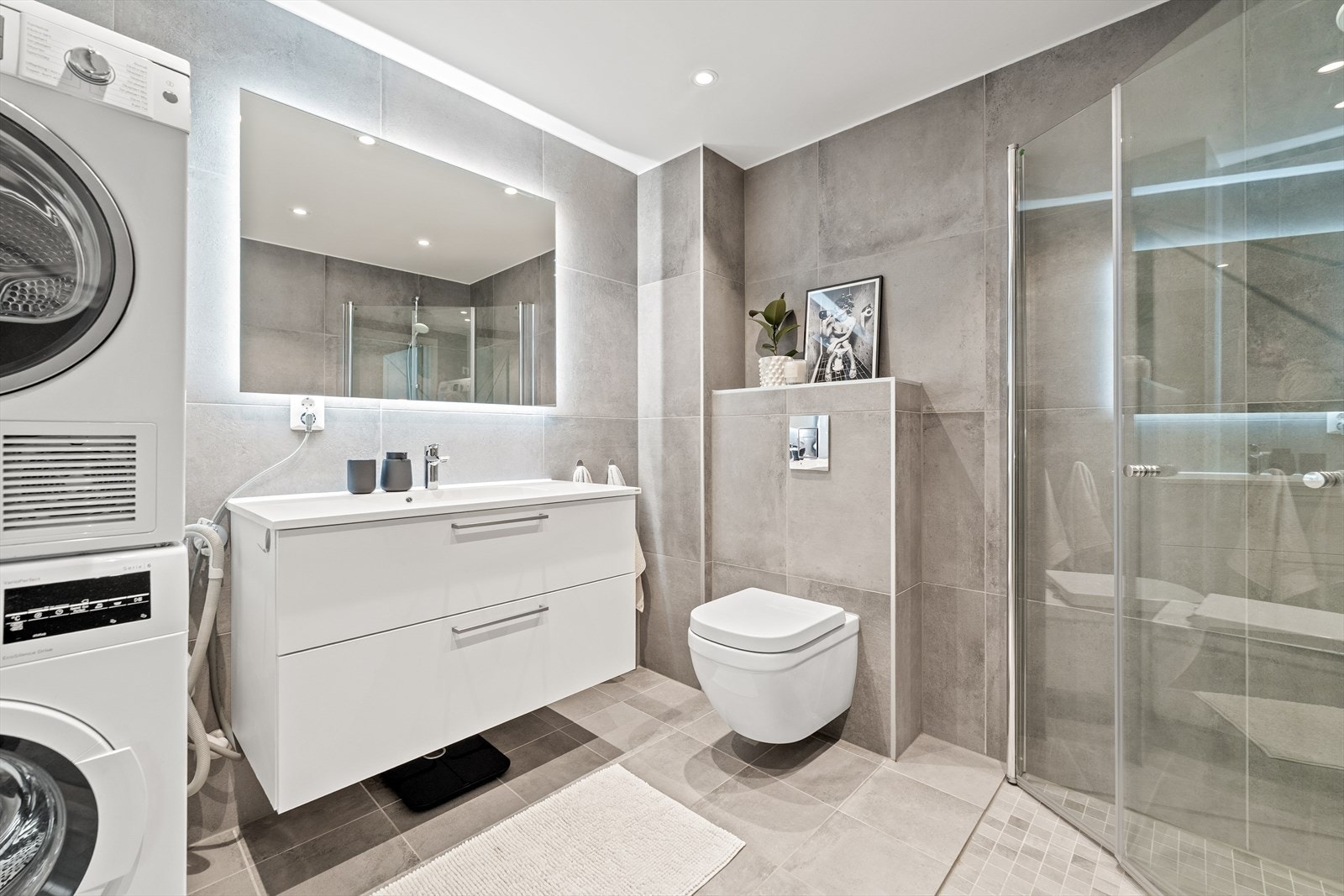 Badet utstyrt med innredning bestående av nedfelt servant med underskap og 1-greps blandebatteri, speil, dusjhjørne med glassdører, vegghengt toalett og opplegg for vaskemaskin. Galleribilde