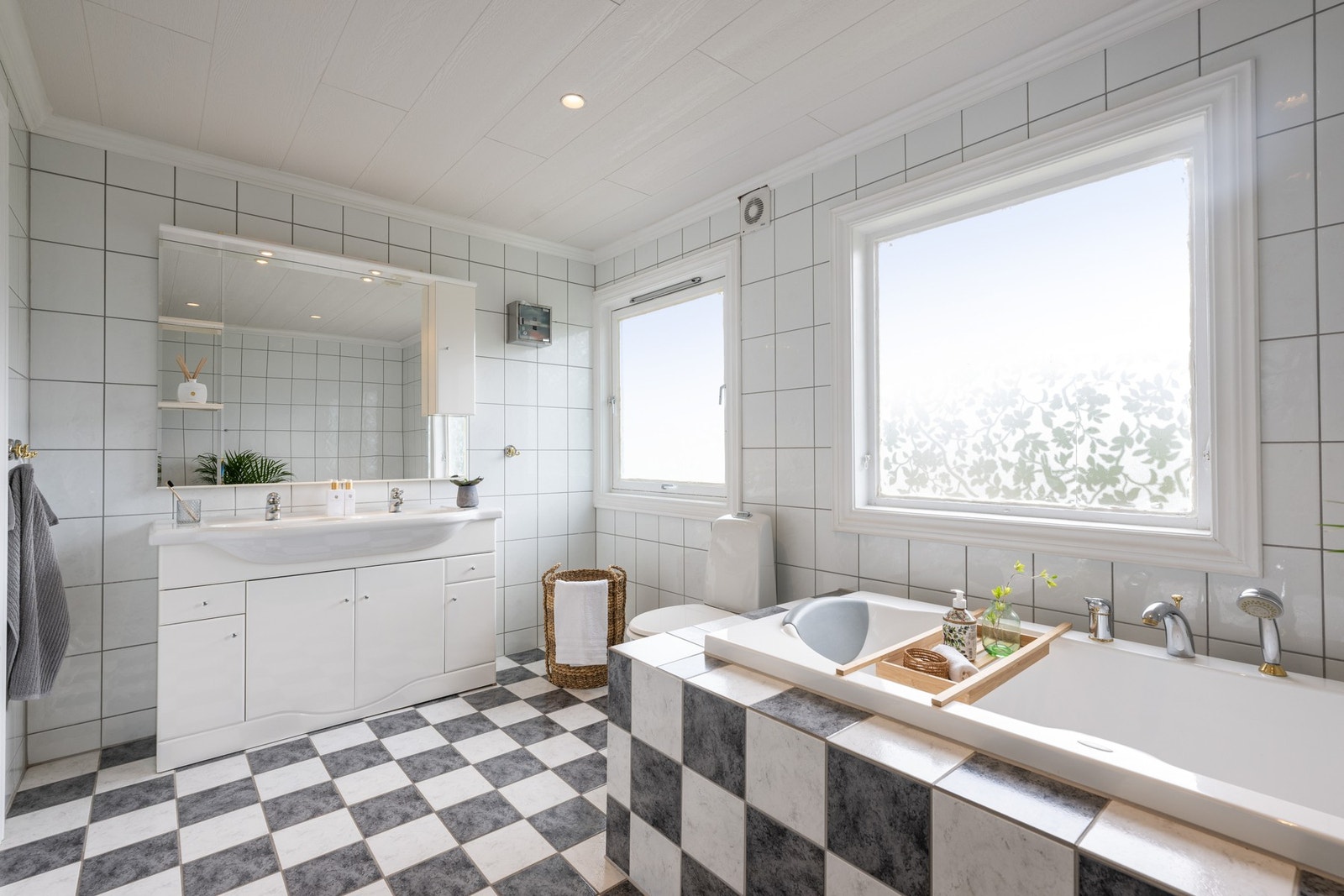Badet har god størrelse, innredet med to vinduer, lysegrå veggfliser og sjakkmønstrede fliser på gulvet. Galleribilde
