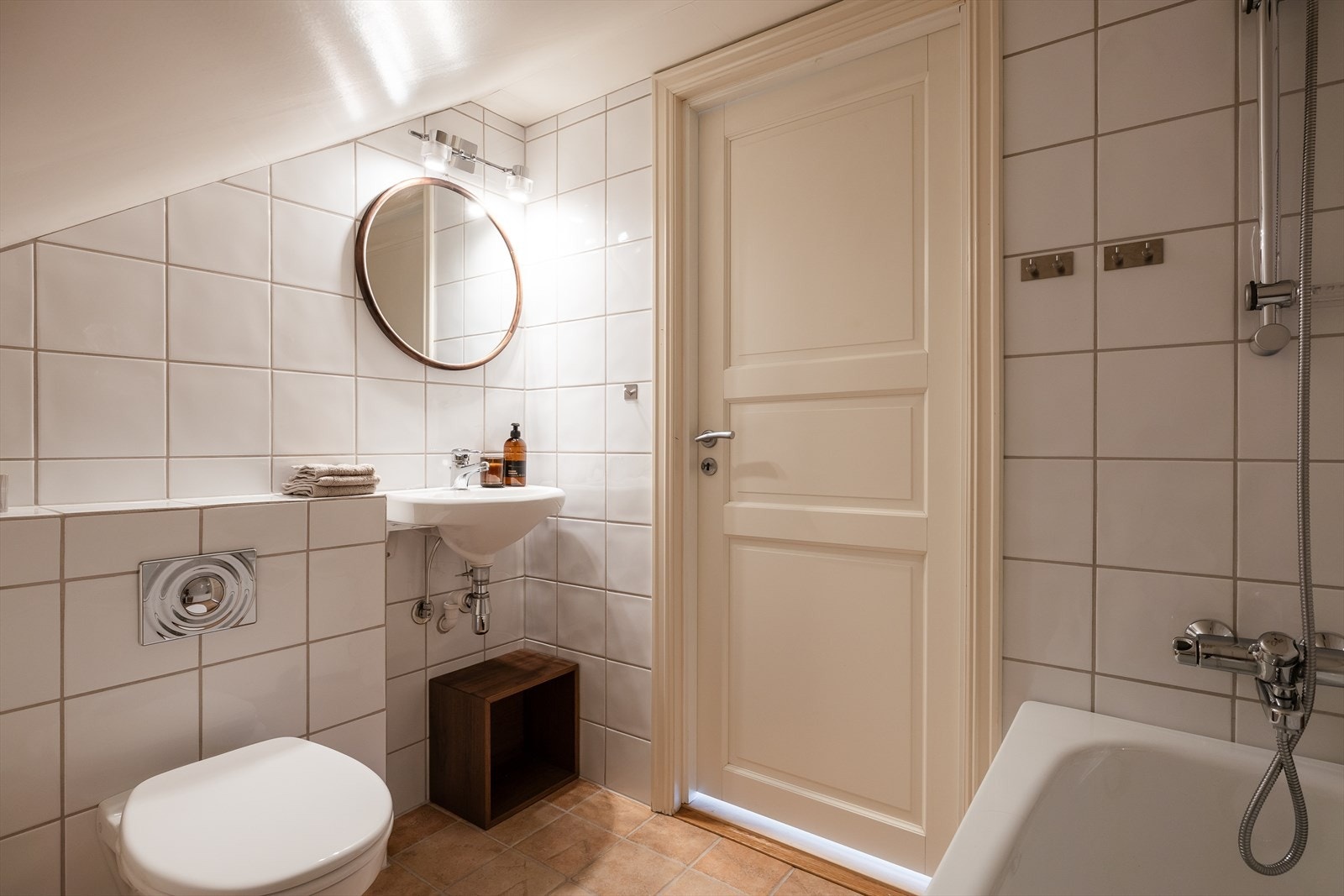 Baderommet er innredet med veggmontert toalett, badekar med dusjbatteri, servant med speil, samt en innkasset varmtvannsbereder. Galleribilde