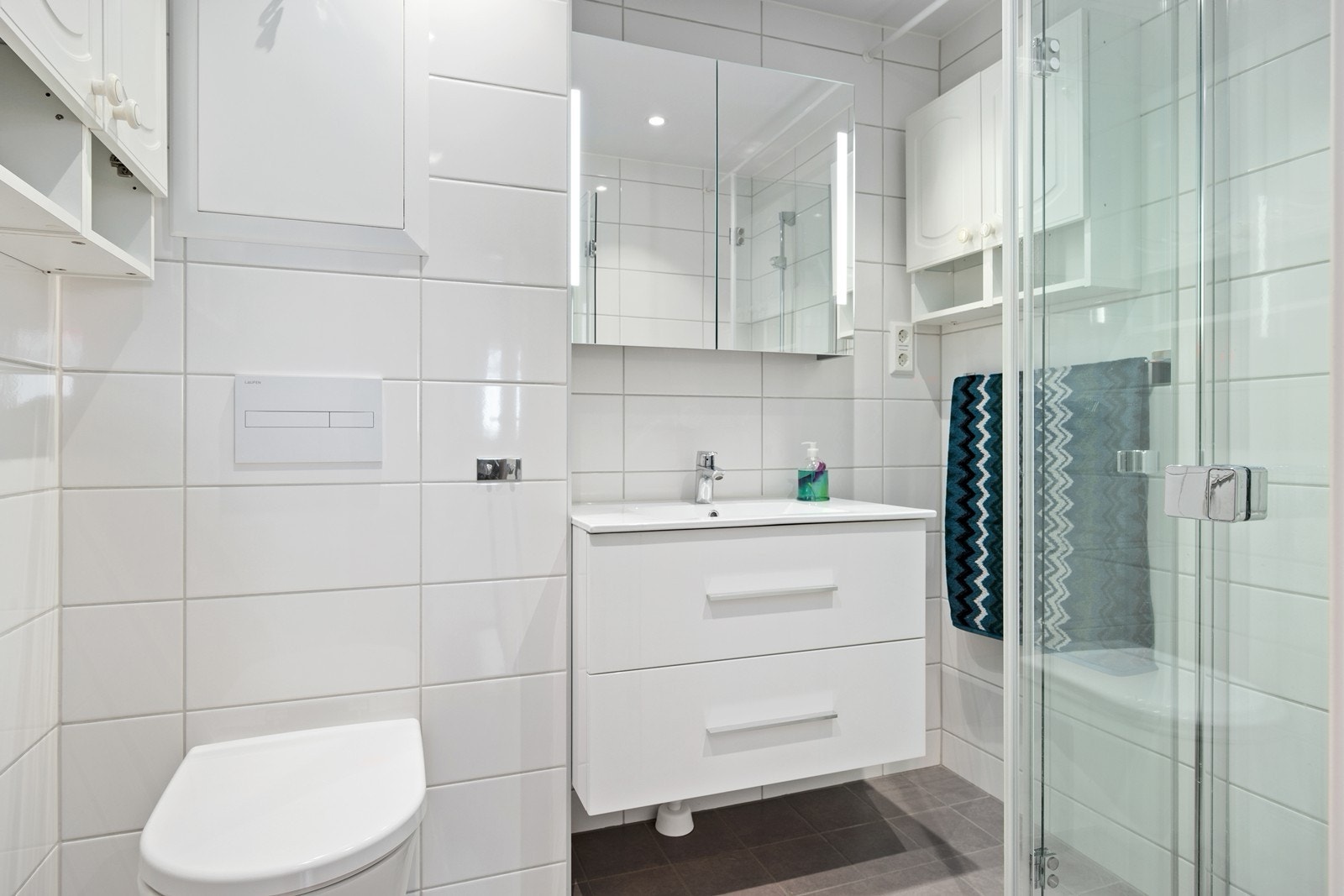 Badet ble oppgradert i regi av borettslaget i 2020. Rommet har grå gulvfliser med gulvvarme, hvite veggfliser og slett hvitt tak med innfelte spotter. Hvit baderomsinnredningen med heldekkende servant, skuffer for oppbevaring og speilskap med belysning. Galleribilde