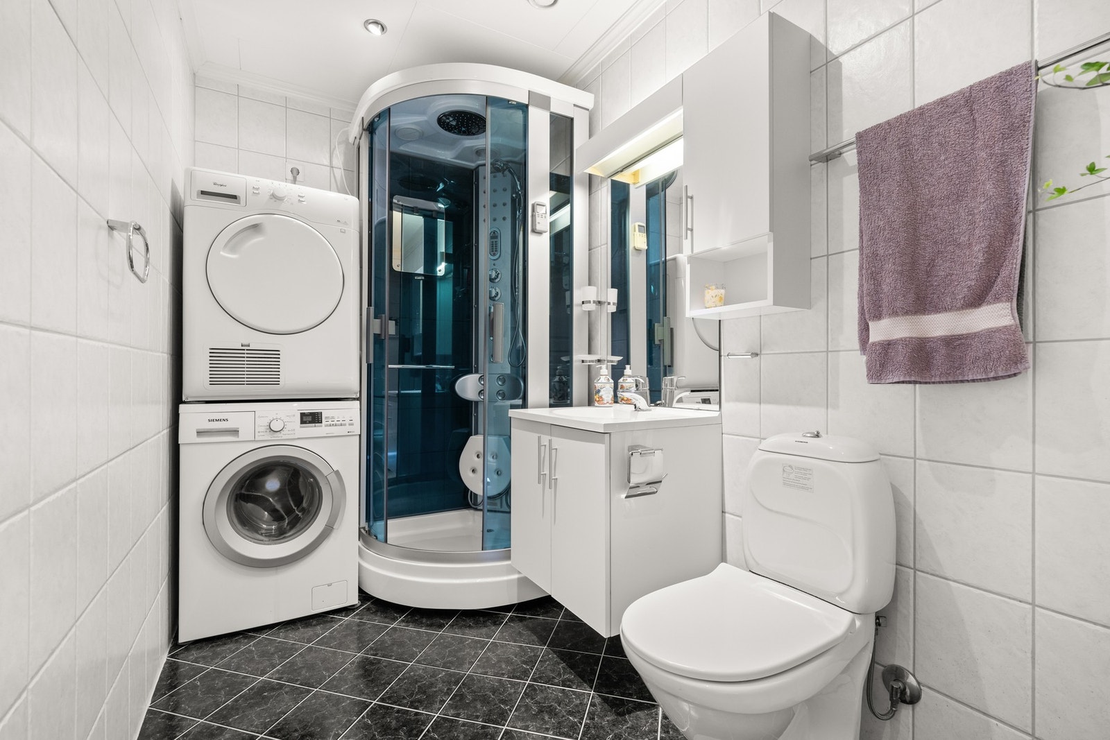 Badet har vegghengt servantskap, dusjarmatur tilkoblet hånddusj og regndusj samt massasjedyser, gulvstående toalett i tillegg til opplegg for vaskemaskin og tørketrommel Galleribilde