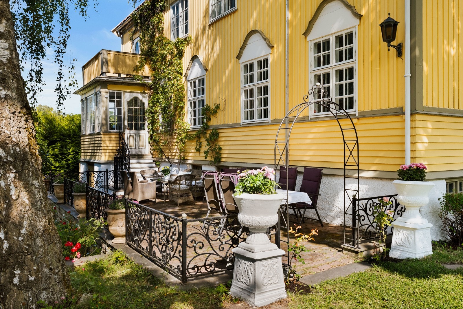 Frodig og inngjerdet tomt med en stor stensatt markterrasse med et påkostet smijernsrekkverk, store plenarealer med frodige og flotte trær, busker og bed. Galleribilde