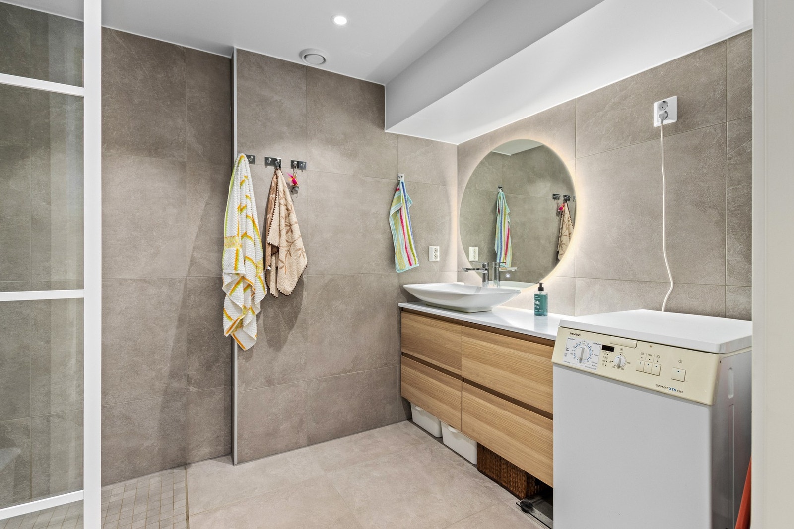 Badet inneholder åpen dusj med glassvegg, vegghengt wc, servant i innredning, opplegg for vaskemaskin og tørketrommel. Galleribilde