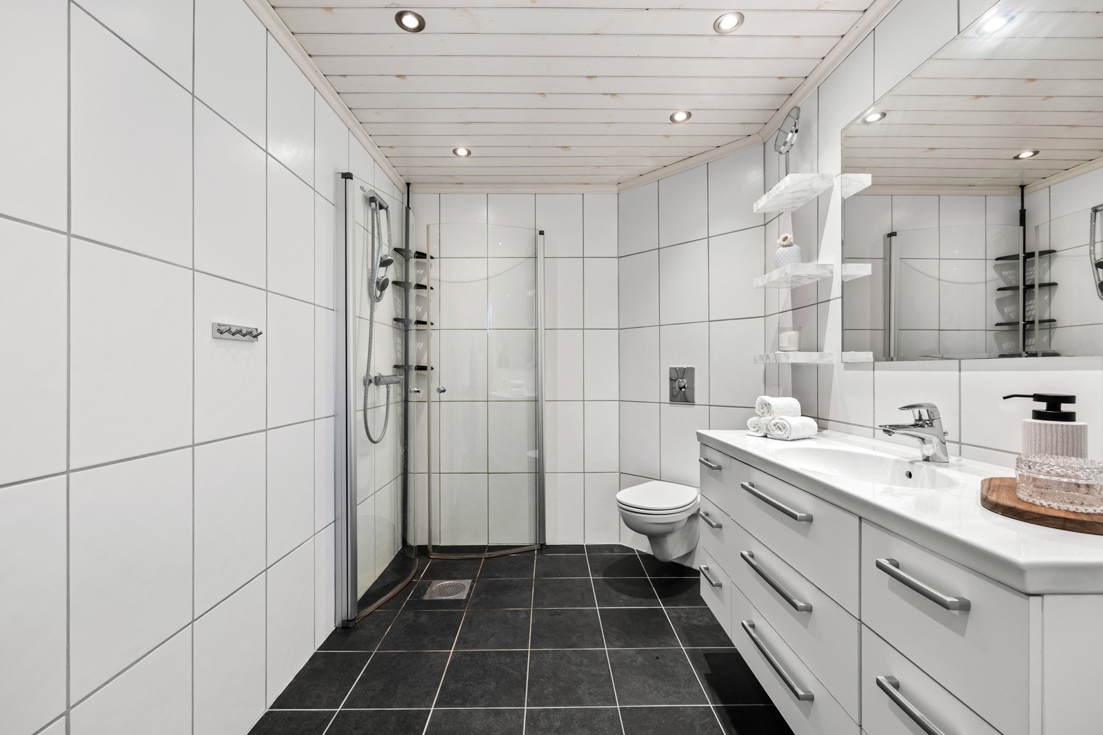 Videre har badet speil, hyller og heldekkende servant på hvitt skuffemøblement, vegghengt toalett og dusjhjørne. Galleribilde