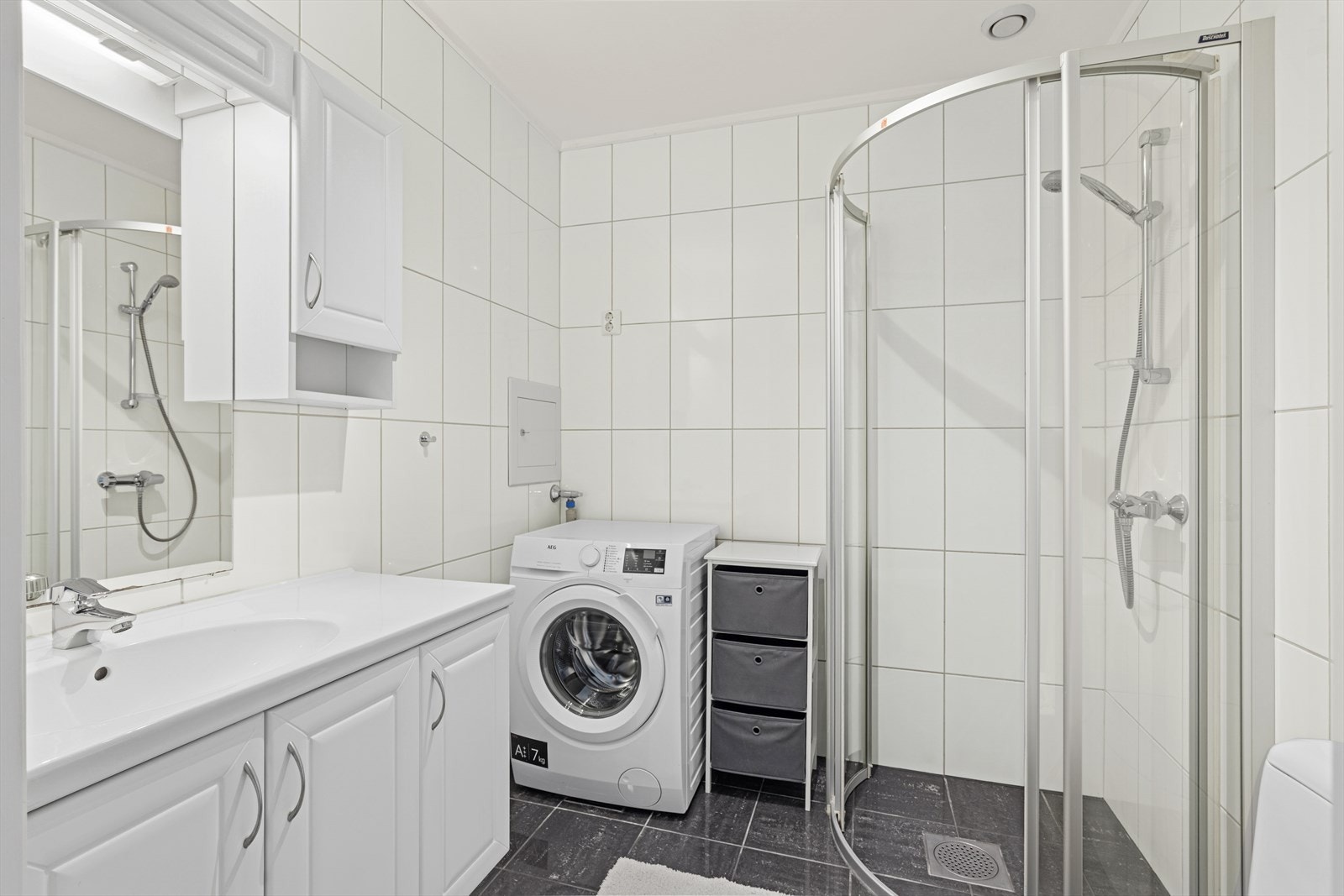 Badet har flis på gulv og vegg, malt gips i tak. Dusjnisje, baderomsinnredning med speil, gulvstående toalett, opplegg for vaskemaskin. Galleribilde