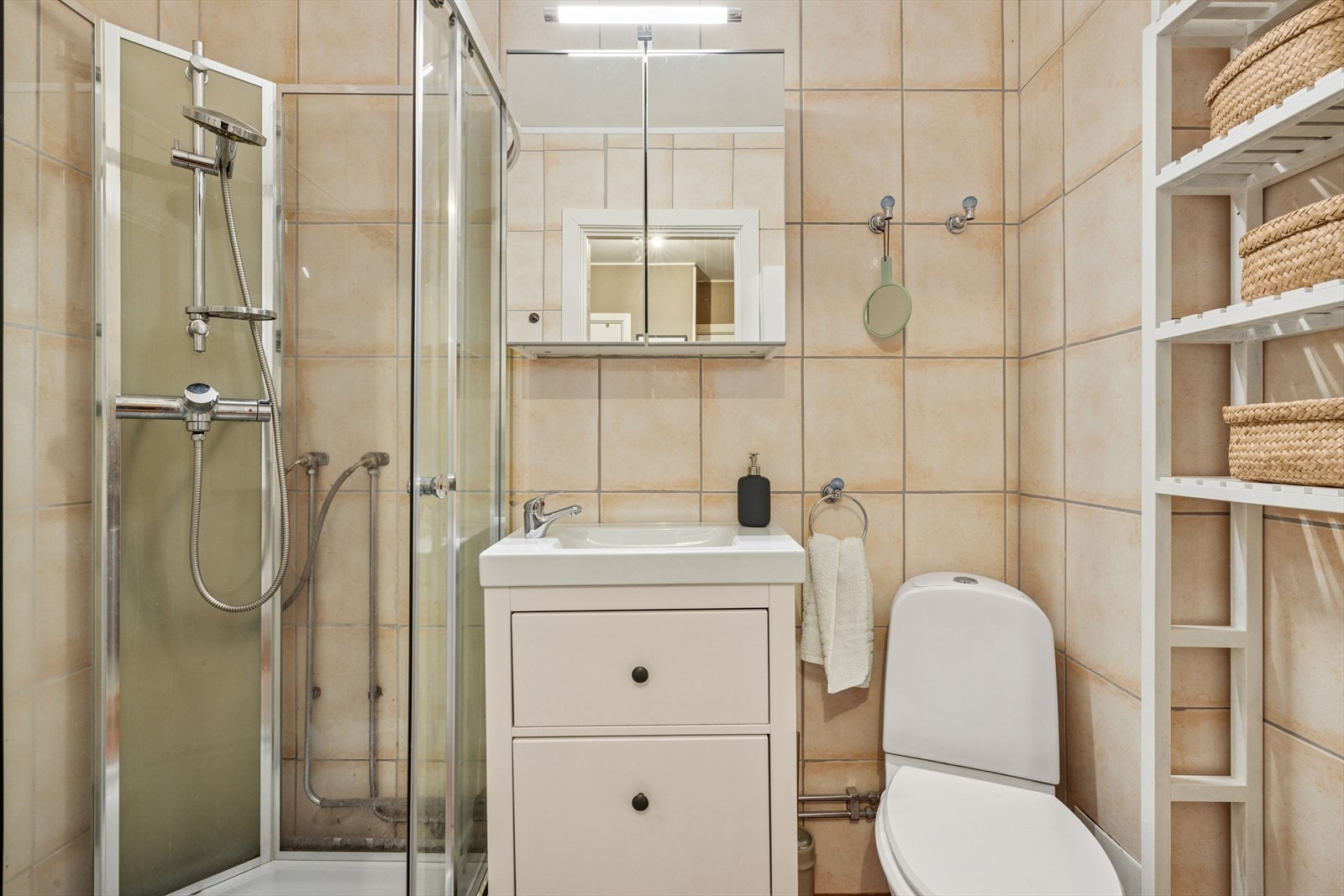 Badet er innredet med servantskap med ett-greps blandebatteri, speilskap, veggskap, gulvstående toalett og dusjkabinett. Galleribilde