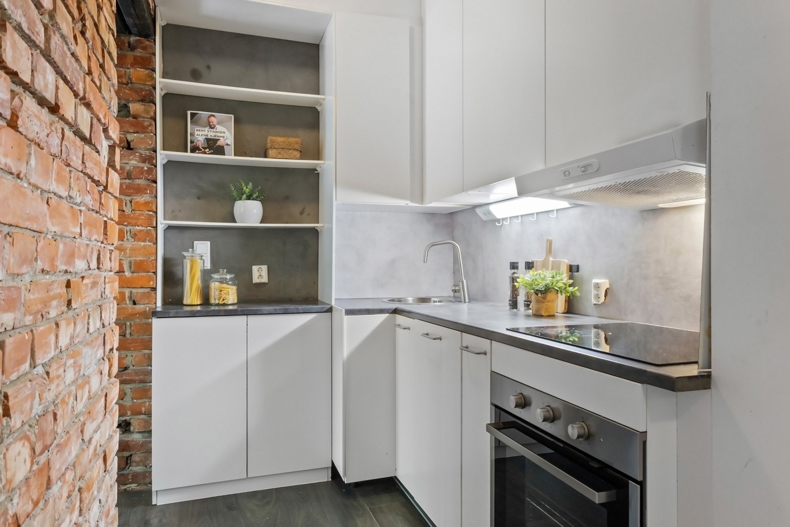 Over benken og i bakkanten av hylle er det grå plater. Kjøkkenet er utstyrt med integrert induksjonstopp, stekeovn og oppvaskmaskin, samt en kullfilterventilator over kokesonen. Galleribilde