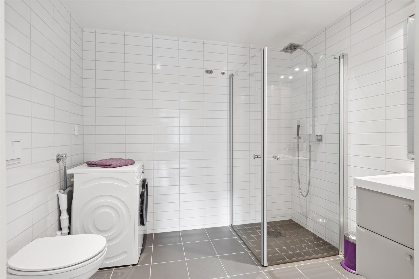 Baderomsinnredningen har lyse fronter og består av en heldekkende servant på en skapseksjon og et speil med belysning. Videre har badet et vegghengt toalett, opplegg for vaskemaskin og et dusjhjørne med innfellbare glassdører. Galleribilde