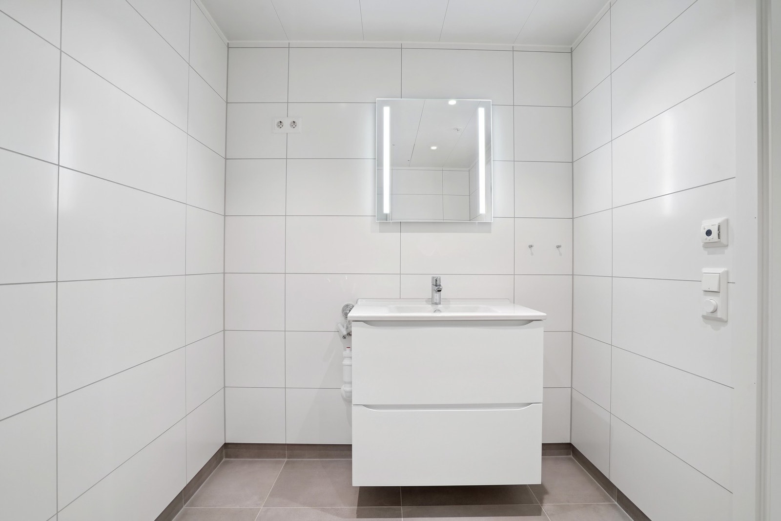 Badet har en stilren utførelse med hvite veggfliser 30x60 cm, grå gulvfliser 60x60 cm. Innredning med hvite skuffer, speil med LED-belysning. Galleribilde