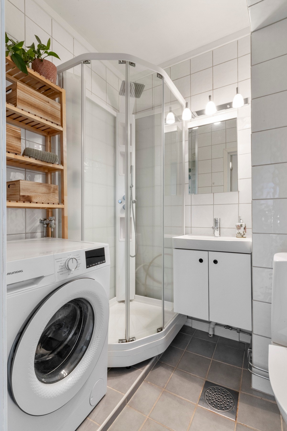 Flislagt baderom med innredning av servant med ett-greps armatur og speil, dusjkabinett med dusjarmatur, frittstående toalett samt opplegg for vaskemaskin. Galleribilde