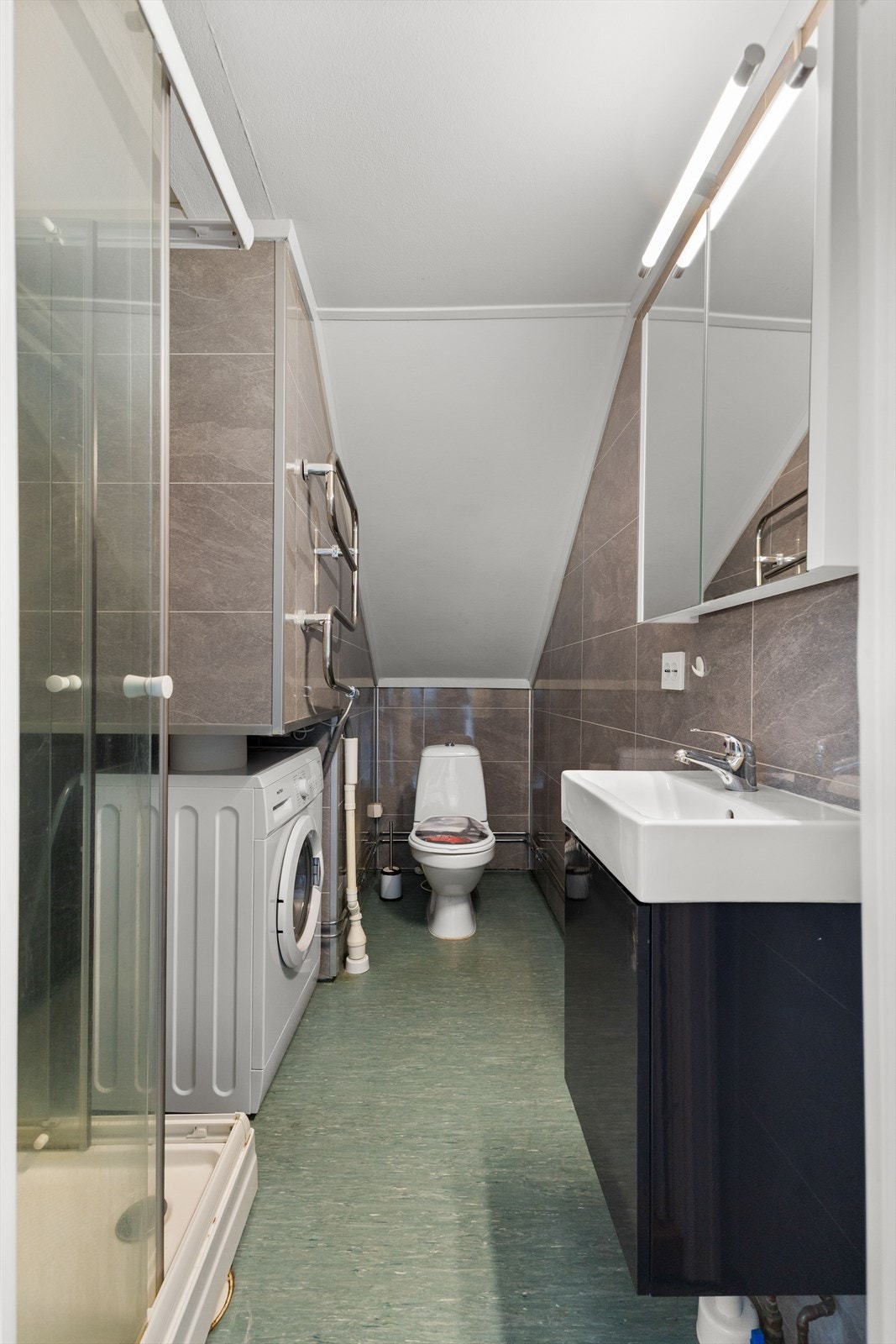 Badet i 2. etasje har baderomsplater på veggene og vinylbelegg på gulvet. Himlingen er kledd med malte plater. Rommet er innredet med dusjkabinett, servant med underskap, toalett og opplegg for vaskemaskin. Galleribilde