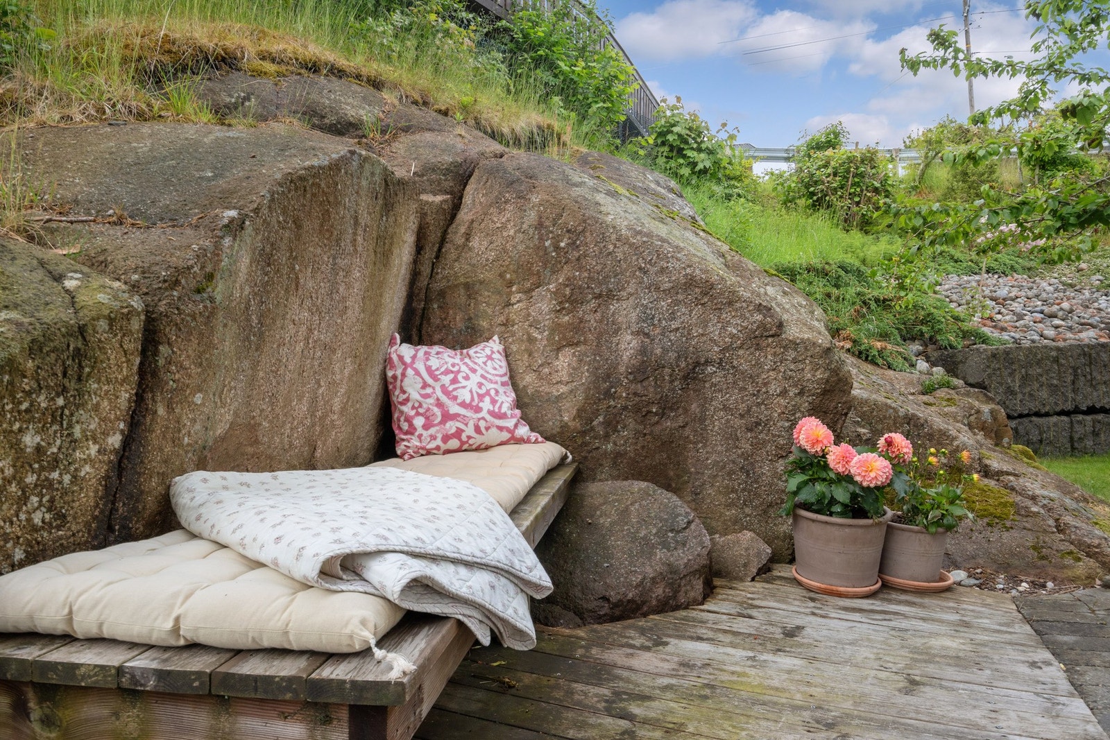 En koselig hage på baksiden, opparbeidet med trær, busker, gressplen og en plassbygd sittebenk langs fine fjellknauser. Galleribilde