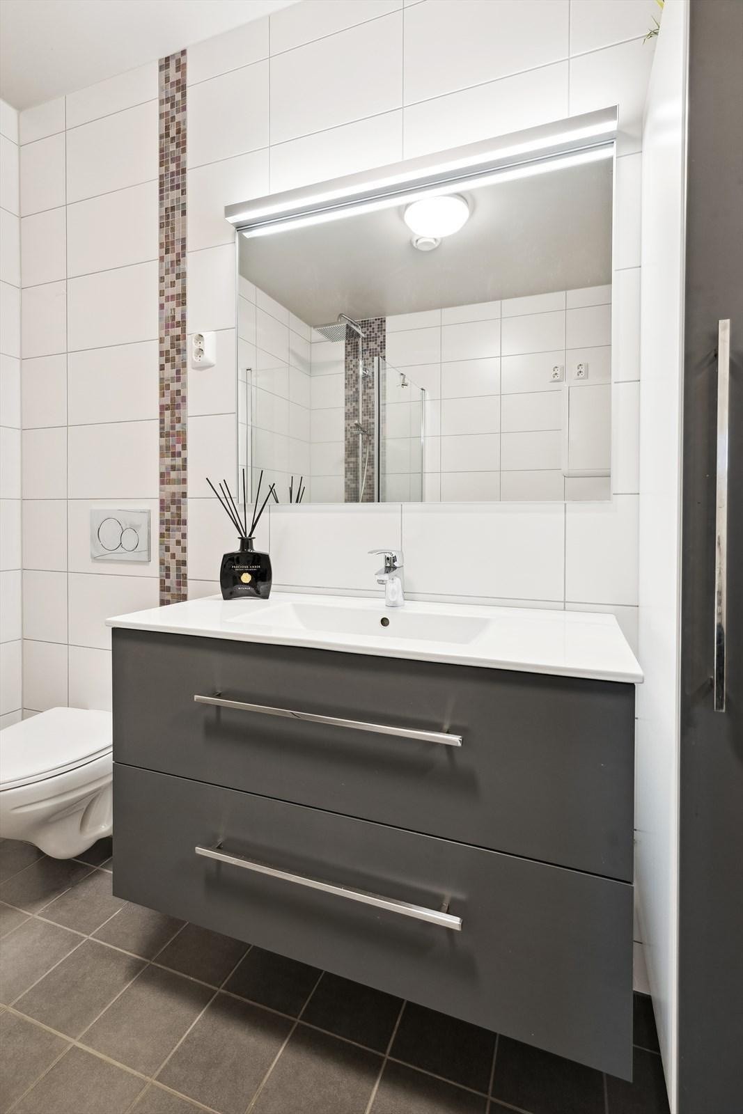 Stilrent flislagt bad med gulvvarme, dusjhjørne med innfellbare glassdører, vegghengt toalett, servant med moderne innredning og opplegg for vaskemaskin Galleribilde