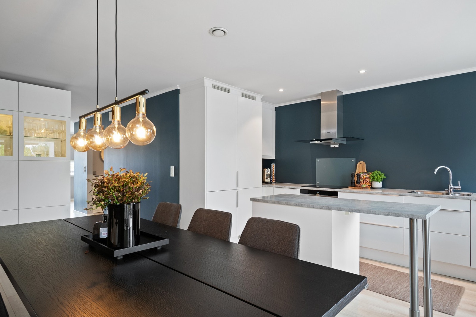 Kjøkkenet har hvite, slette fronter, grå laminatbenkeplater og er montert i L-form med kjøkkenøy og vitrineskap ved spiseplassen. Galleribilde