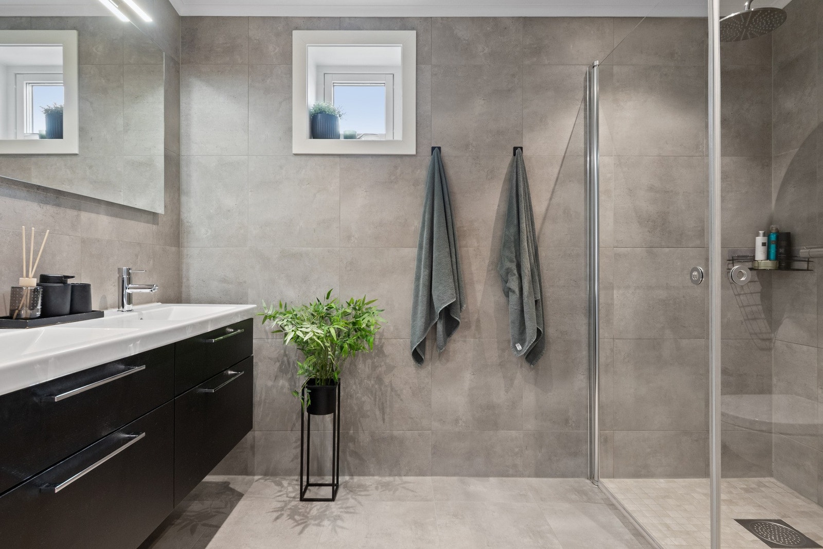 Badet i andre etasje har ekstra takhøyde, flislagte overflater, regnfallsdusj og dobbelservant på sort skuffemøblement. Galleribilde