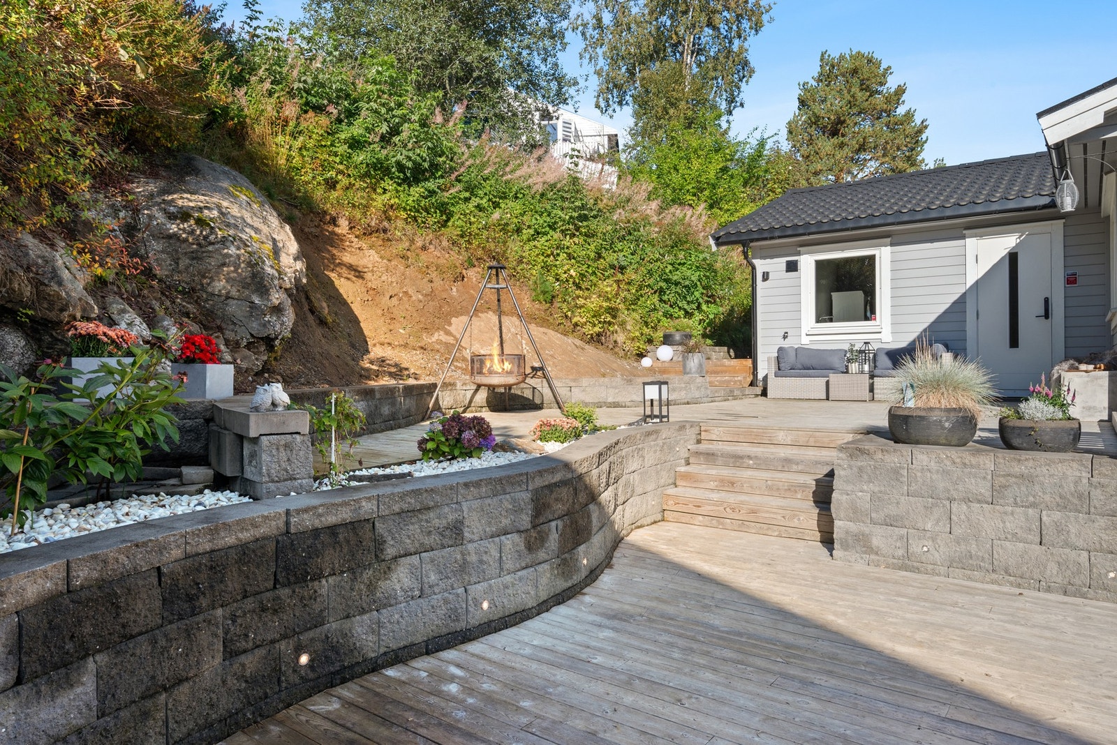 Terrassen har en buet avslutning mot terrenget, med singel og blomster i bed på støttemuren. Galleribilde