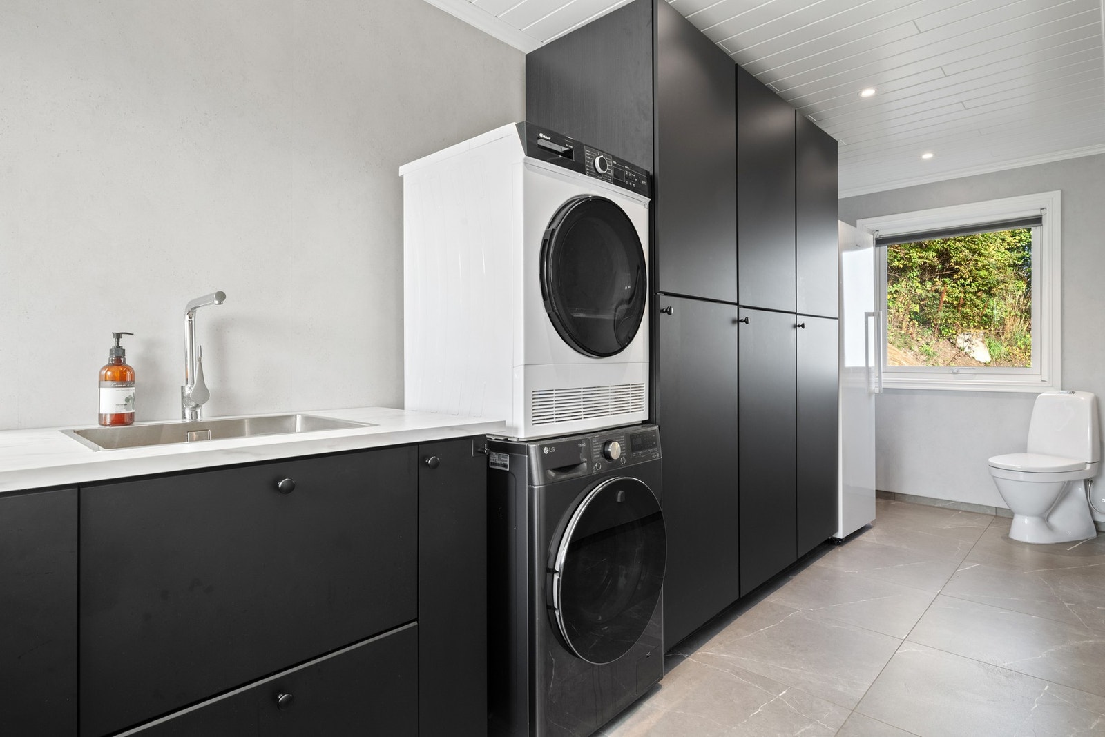 Gulvflisene fra gangen fortsetter inn på et vaskerom fra 2021, utstyrt med store høyskap og et praktisk gjestetoalett. Galleribilde