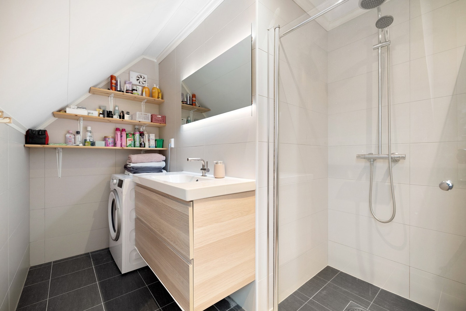 Badet er innredet med vegghengt toalett, dusj med glass-foldedører og baderomsmøbel med speil og belysning. Galleribilde