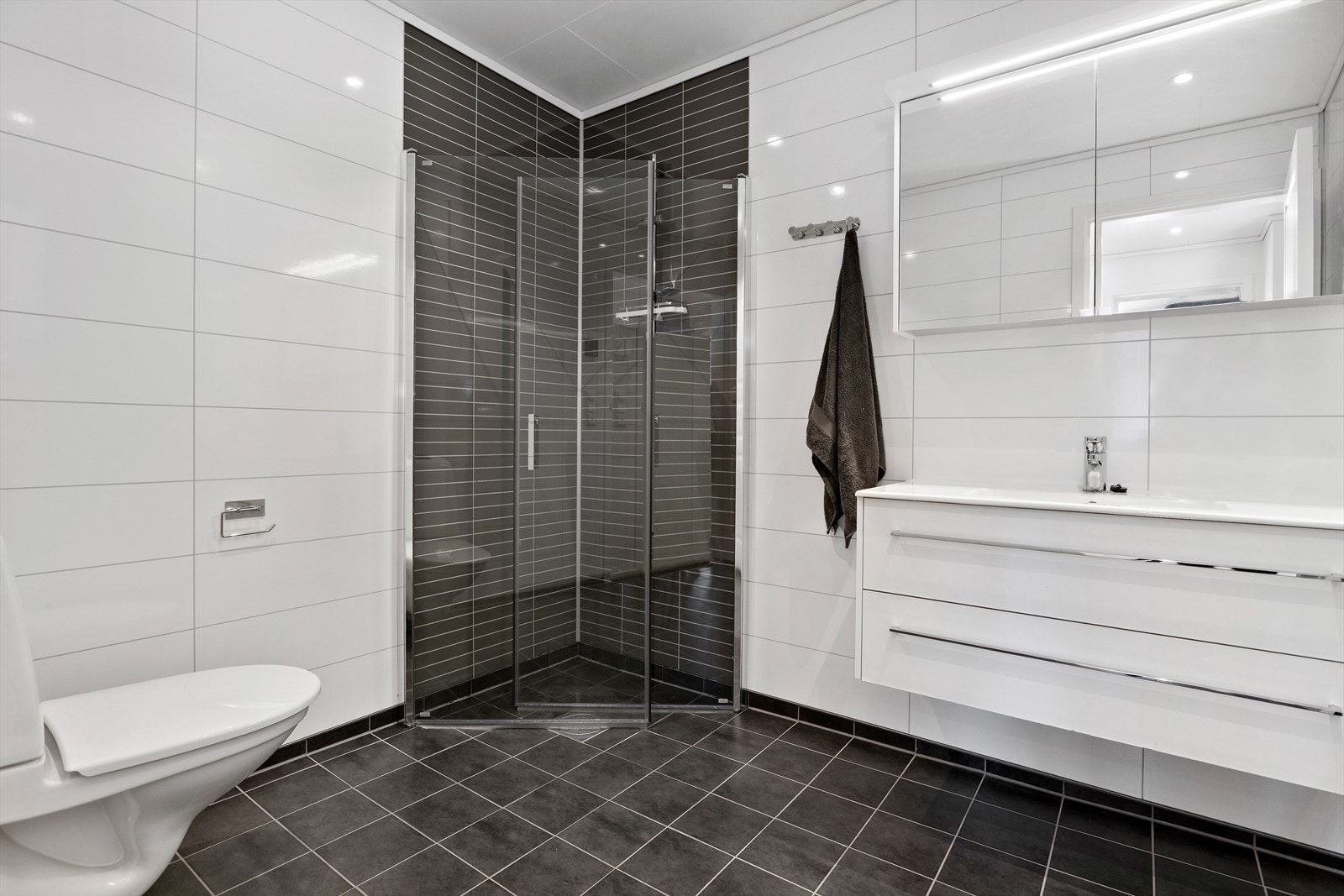 Badet har flislagt gulv med varmekabler. Her er det innredet med servant i underskap, speilskap med lysarmatur, dusjhjørne og vegghengt toalett. Galleribilde