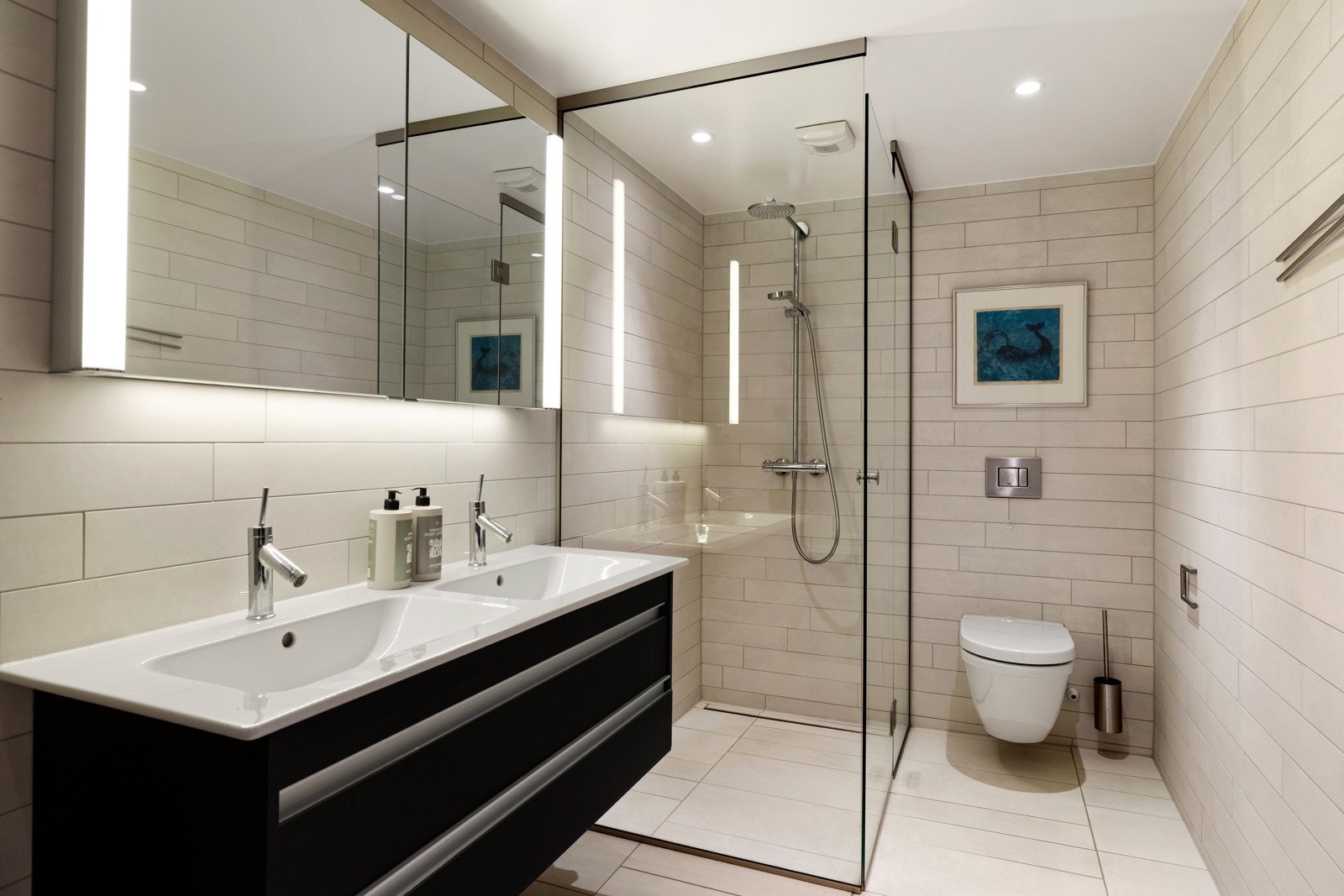 Badet er utstyrt med både badekar og et romslig dusjhjørne med regnfallsdusj og glassdør. Videre har badet vegghengt wc, dobbelservant med praktisk skuffeløsning, samt et stort speilskap med integrert belysning Galleribilde