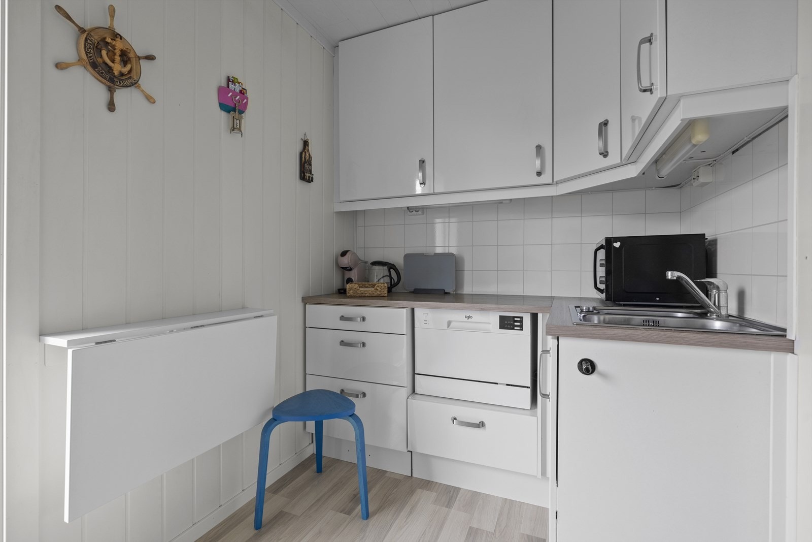 Kjøkkeninnredningen består av hvite, glatte fronter, benkeplate i laminat med nedfelt kum i rustfritt stål, belysning under overskap og kjøkkenplater på vegg over benkeskap. Galleribilde