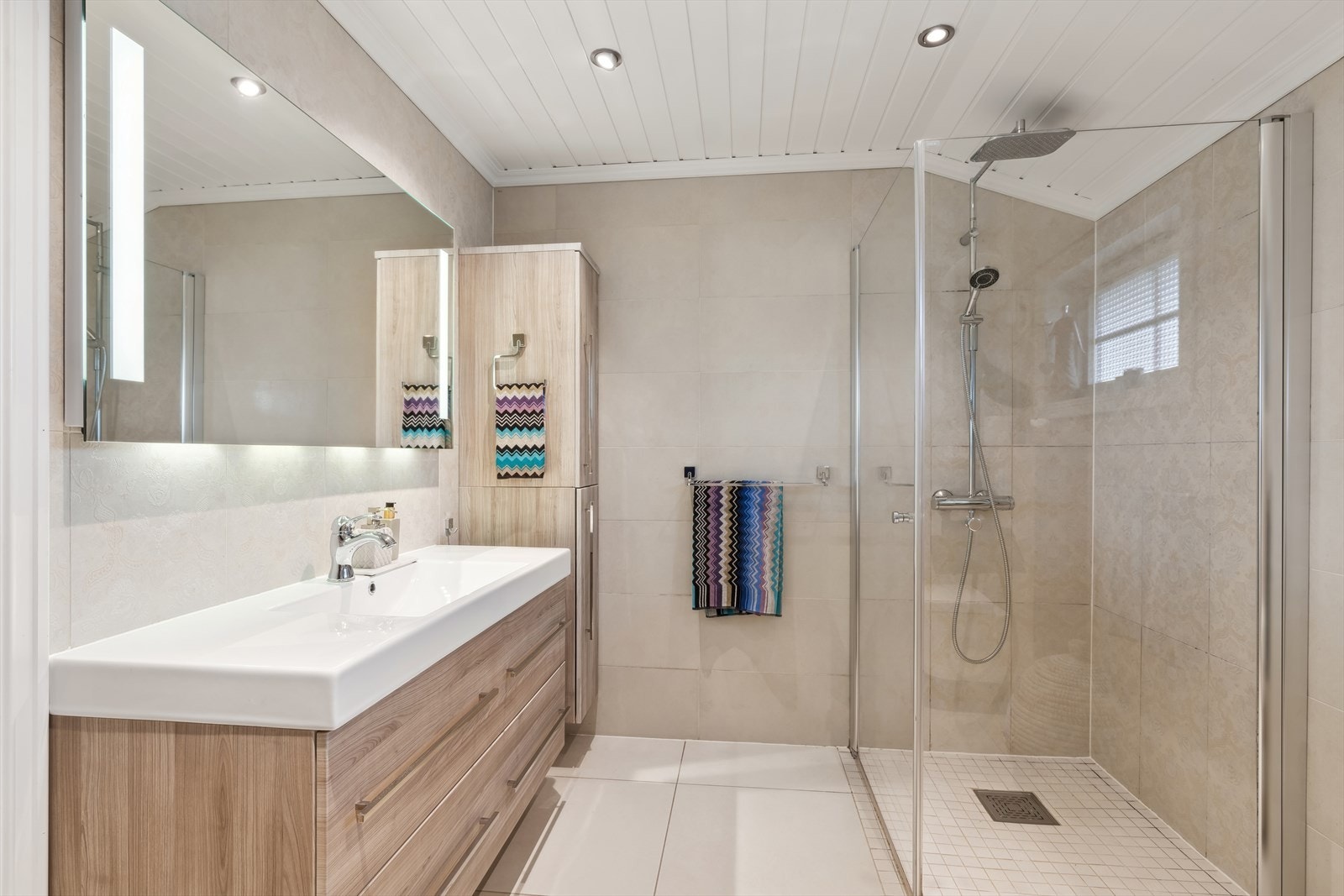 Rommet er innredet med en stor baderomsinnredning med servant og skuffseksjon, høyskap montert på vegg, vegghengt toalett og en stor og romslig dusjsone som er pent gjennomført med regnfallsdusj og innfellbare glassdører. Galleribilde