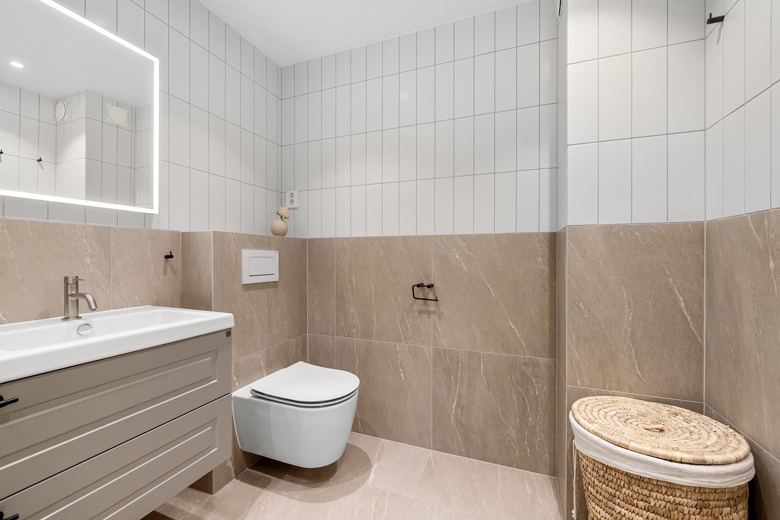 Badet inneholder dusjhjørne med glassdør og veggnisje, regnfallsdusj og hånddusj, vegghengt toalett og baderomsrinnredning med skuffer, dobbel servant med to ett-greps blandebatterier og belyst speil på vegg. Galleribilde