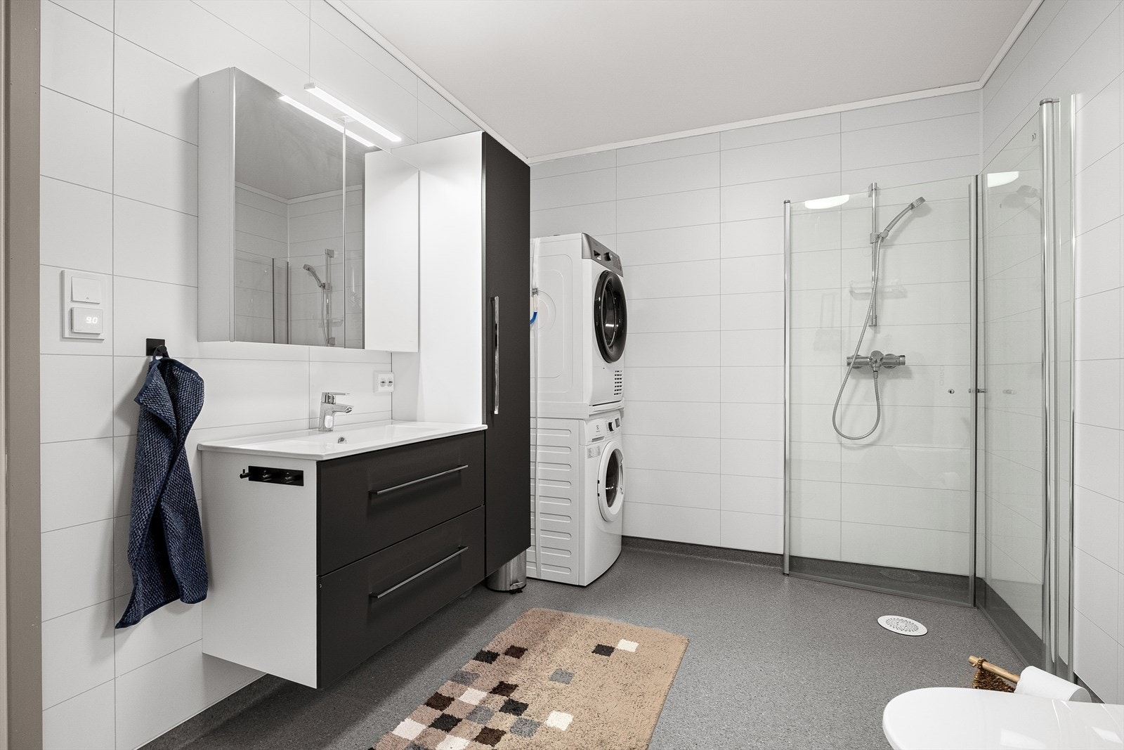 Flott og lyst bad som inneholder vegghengt toalett, dusjhjøre, servant med underskap og opplegg for vaskemaskin. Galleribilde