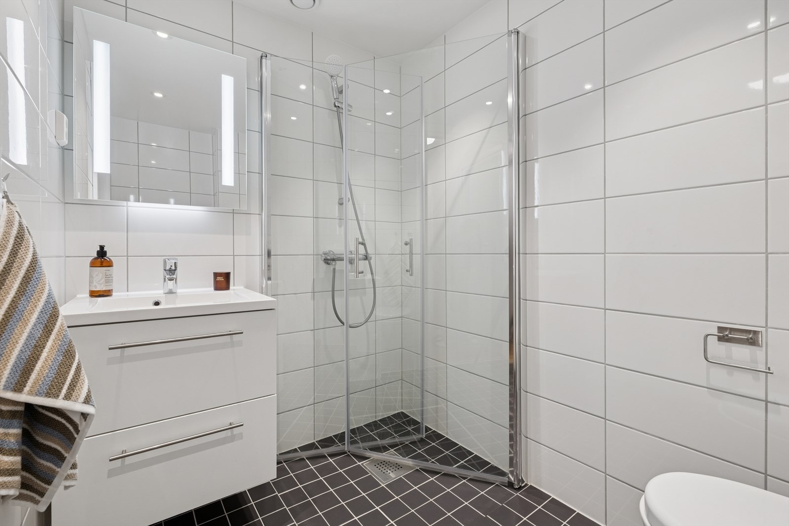 Pent flislagt baderom med vannbåren gulvvarme som er innredet med vegghengt klosett, servant med innredning, speil med lys og dusjhjørne med svingbare glassdører. Det er balansert ventilasjon med avtrekksventil i himling. Galleribilde
