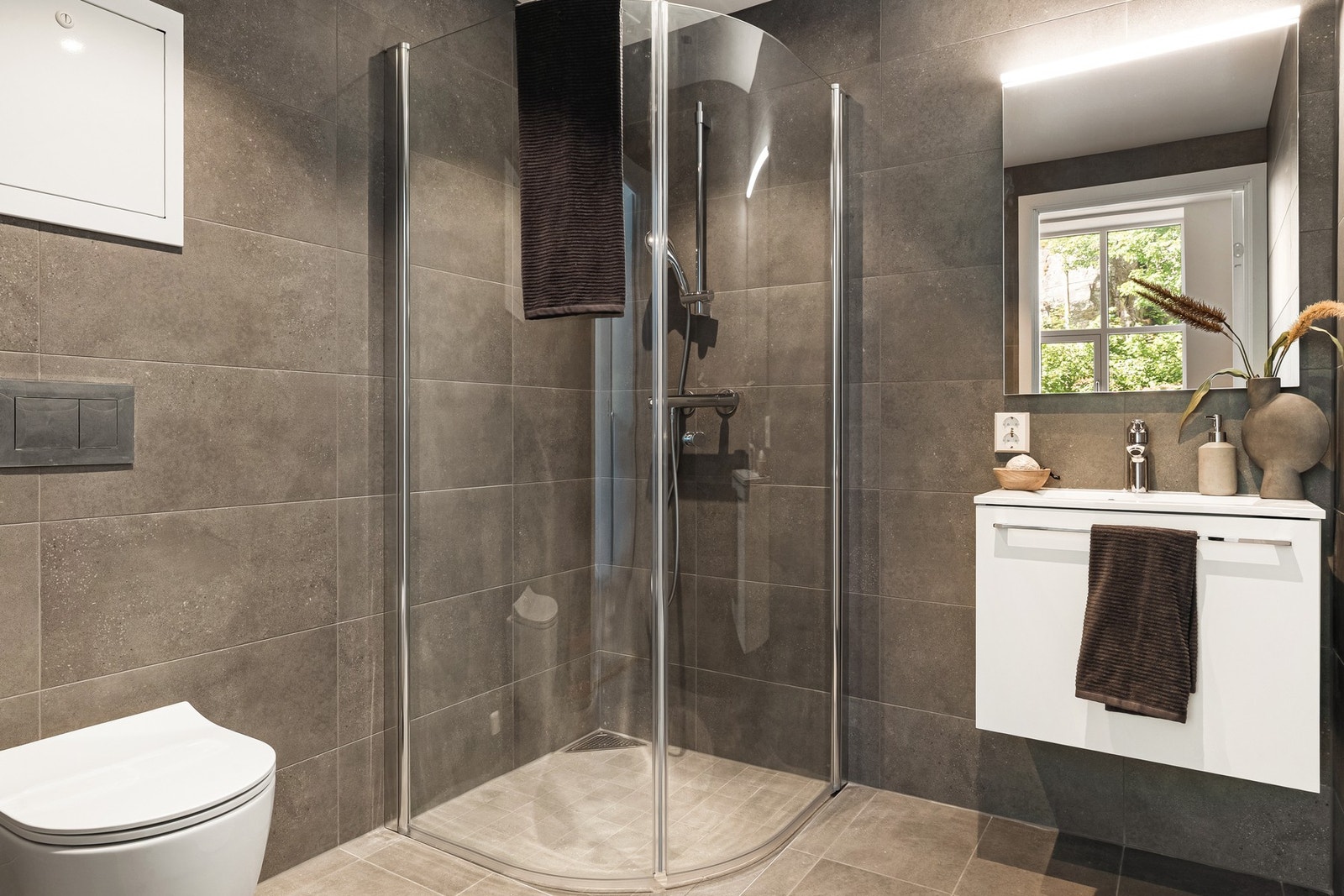 Lekkert bad i grå fliser med varme i gulvet, innredet med dusjhjørne, vegghengt toalett og servant med møblement og belysning. Galleribilde