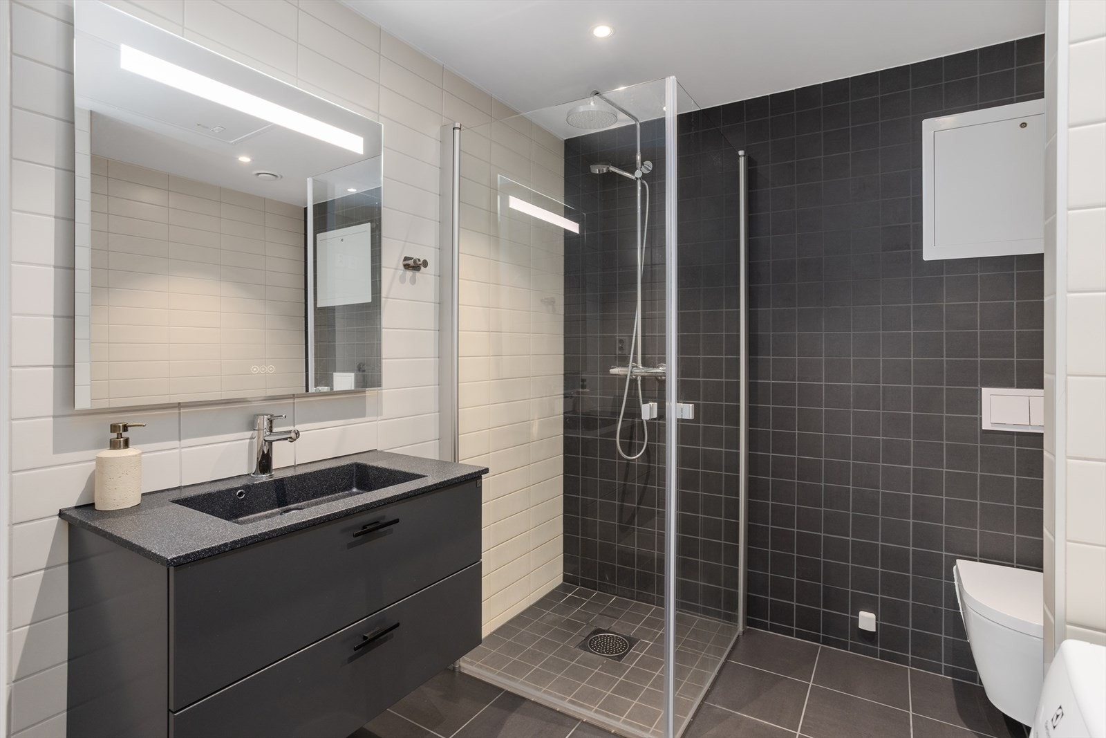 Kombinert bade-/vaskerom med fliser på gulv (gulvvarme ) og vegg. Innredet med veggmontert wc, nedsenket vask med innredning og speil, nedsenket dusjnisje med glassdører og opplegg til vaskemaskin/tørketrommel. Galleribilde