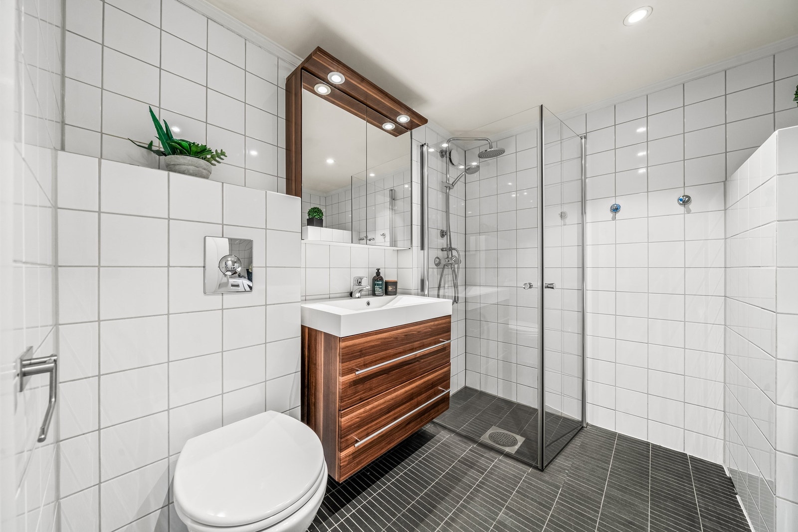 Badet er utstyrt med dusjhjørne med glassvegger og dusj direkte på gulv med termostatstyrt armatur, veggmontert toalett, servantskap med skuffer og speilskap med belysning. Galleribilde