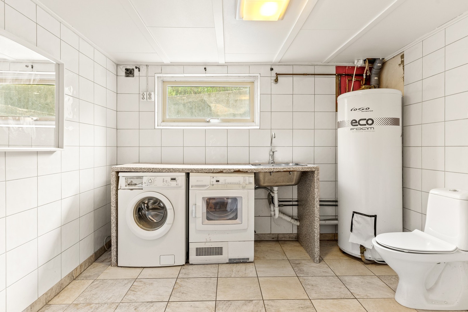 Vaskerom med opplegg for vaskemaskin, tørketrommel, varmtvannsbereder og toalett. Galleribilde