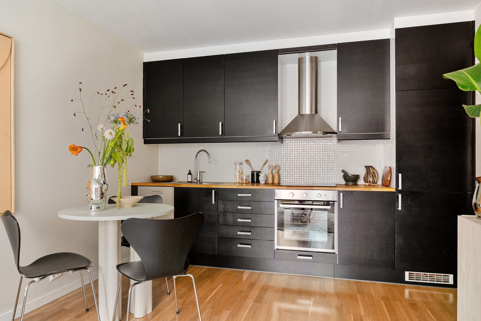 Kjøkken innredet med mørke, slette fronter med stålhåndtak, overskap med benkebelysning og lyse mosaikkfliser på vegg Galleribilde