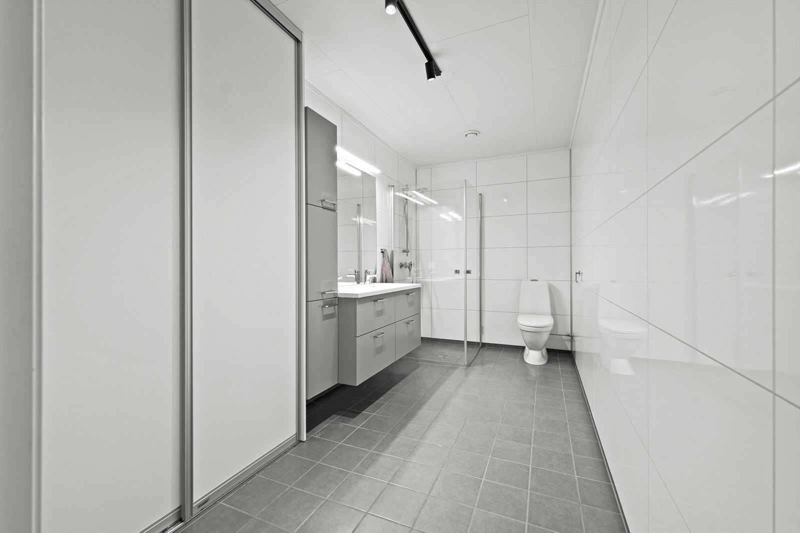 Pent og tidløst bad med fliser på gulv og baderomsplater på vegger. Den smarte skyvedørsgarderoben gjemmer vaskemaskin. Galleribilde