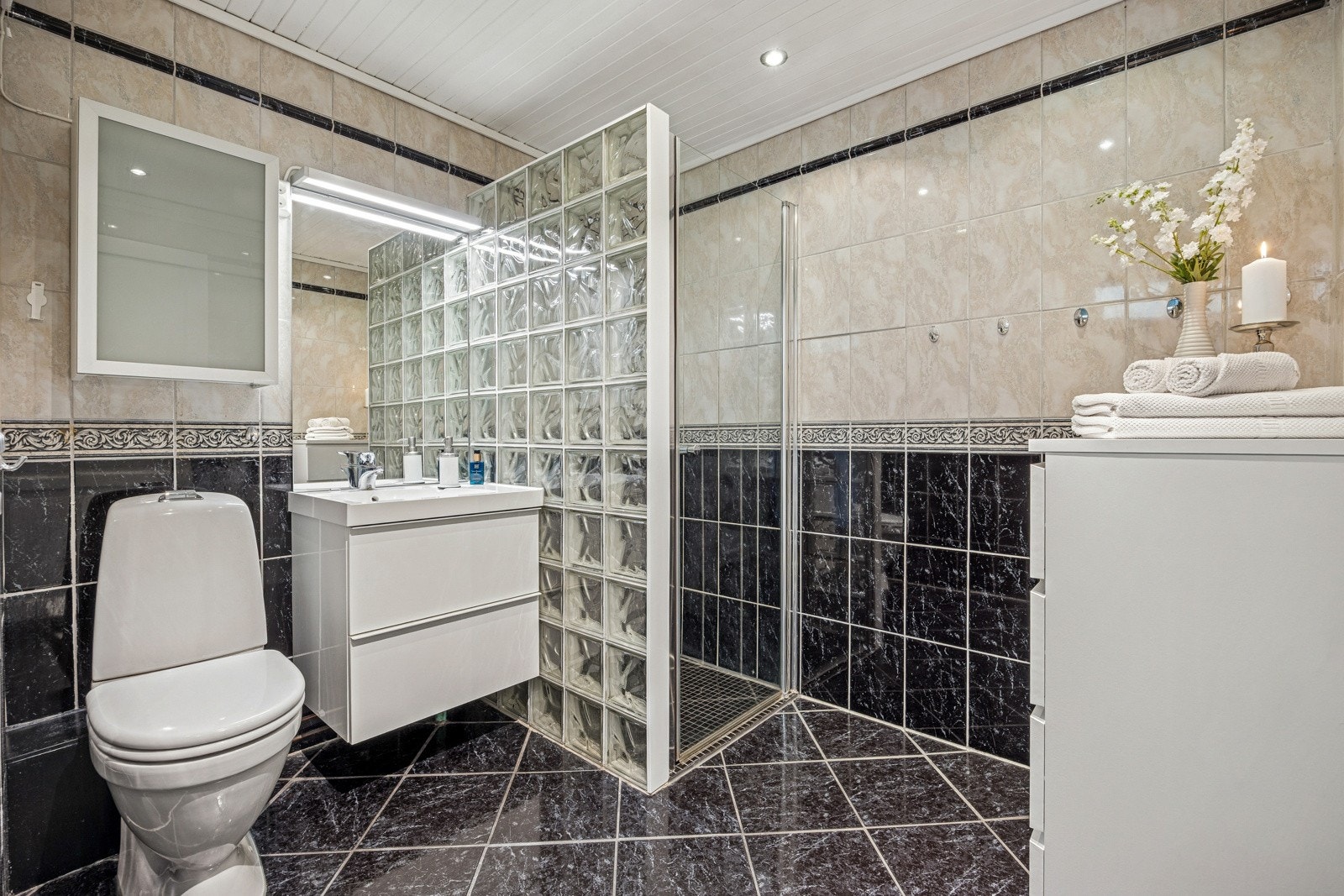 Pent flislagt bad med toalett, dusjnisje og en lys innredning med skuffer og nedfelt servant. Galleribilde