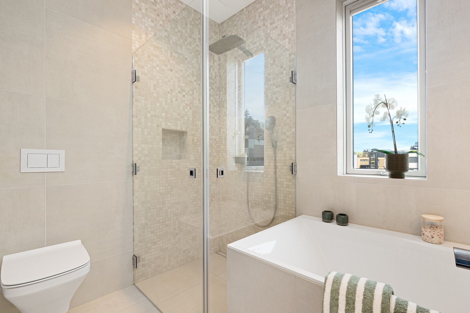 Badet er utstyrt med hvit skuffeinnredning med helstøpt servantbenk, speil på vegg med tilhørende lyspunkt, plassbygd badekar, dusjhjørne med innfellbare glassvegger og vegghengt toalett. Galleribilde