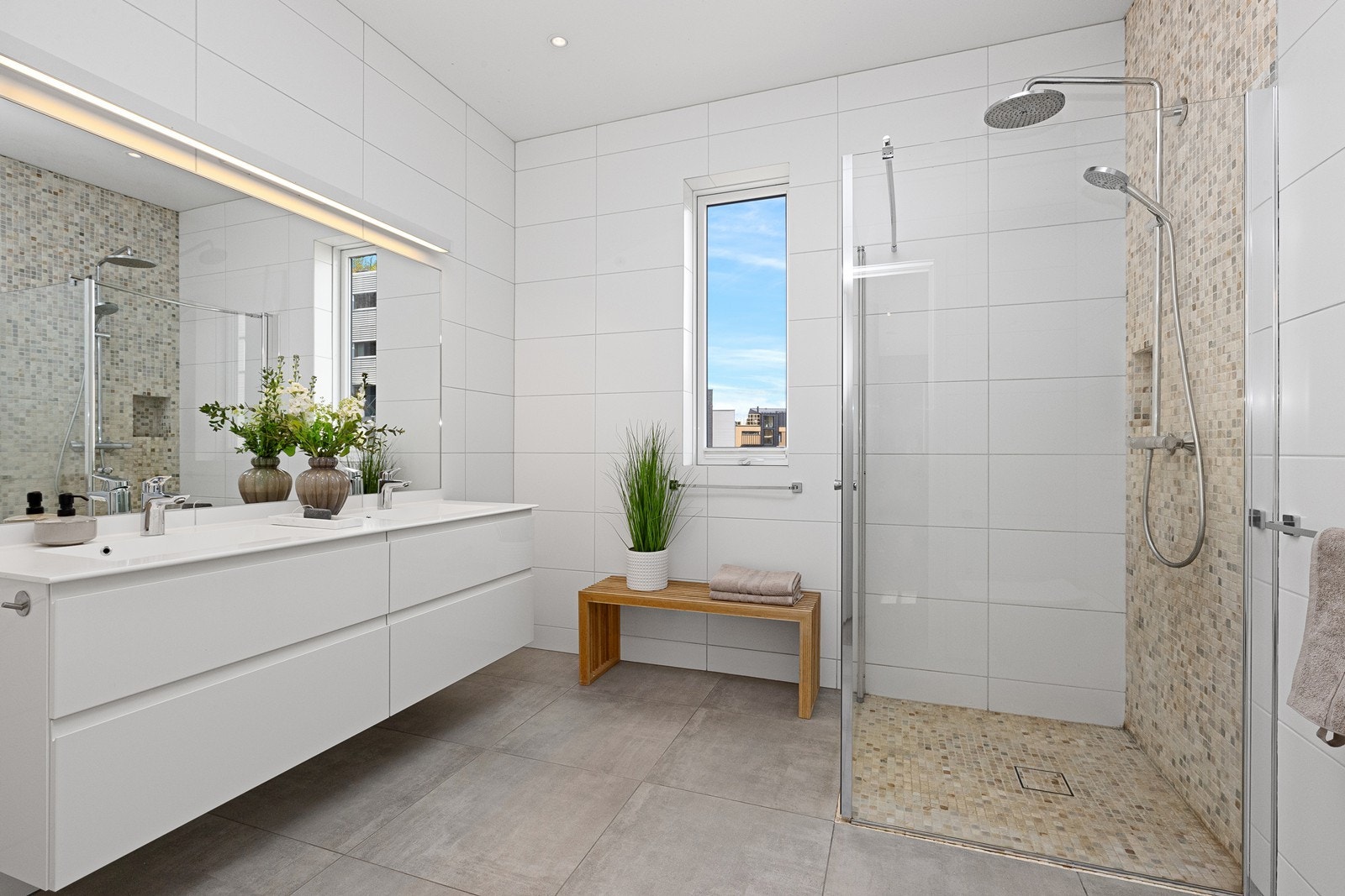 Bad 1. etasje utstyrt med hvit skuffeinnredning med helstøpt dobbel servantbenk, speil på vegg med lysskinne, dusjhjørne med glassvegger og vegghengt toalett. Galleribilde